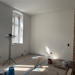 Denis Tar - Pokój po malowaniu ścian na biało z widocznymi narzędziami malarskimi, zabezpieczonymi oknami i fragmentem ściany z cegły. Podłoga częściowo zabezpieczona folią.