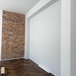 Denis Tar - Pokój po remoncie z białą ścianą, fragmentem ściany z cegły i parkietem w jodełkę. Widoczne narzędzia i materiały wykończeniowe.