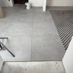 Denis Tar - Ułożone szare płytki podłogowe w dużym formacie, kontrastujące z fragmentem podłogi w czarno-białą szachownicę. Widoczny statyw i wiadro z farbą.