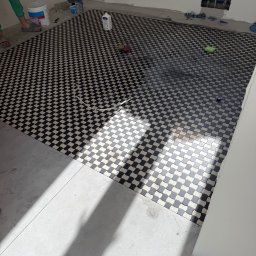 Denis Tar - Ułożona podłoga z czarno-białej mozaiki w łazience, widoczne narzędzia glazurnicze i resztki materiałów budowlanych. Częściowo ułożone płytki podłogowe.