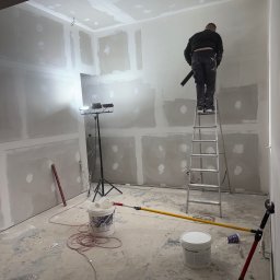 Denis Tar - Prace wykończeniowe w nowym mieszkaniu: mężczyzna na drabinie szpachluje ścianę z płyt gipsowo-kartonowych, widoczne narzędzia, oświetlenie i wiaderka z masą szpachlową.
