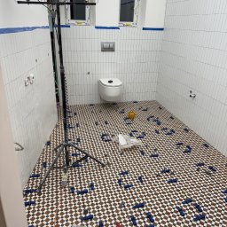 Denis Tar - Układanie mozaiki w łazience w Chorzowie. Na podłodze brązowo-biała szachownica z niebieskimi klinami poziomującymi. Ściany wyłożone białymi, pionowymi płytkami.