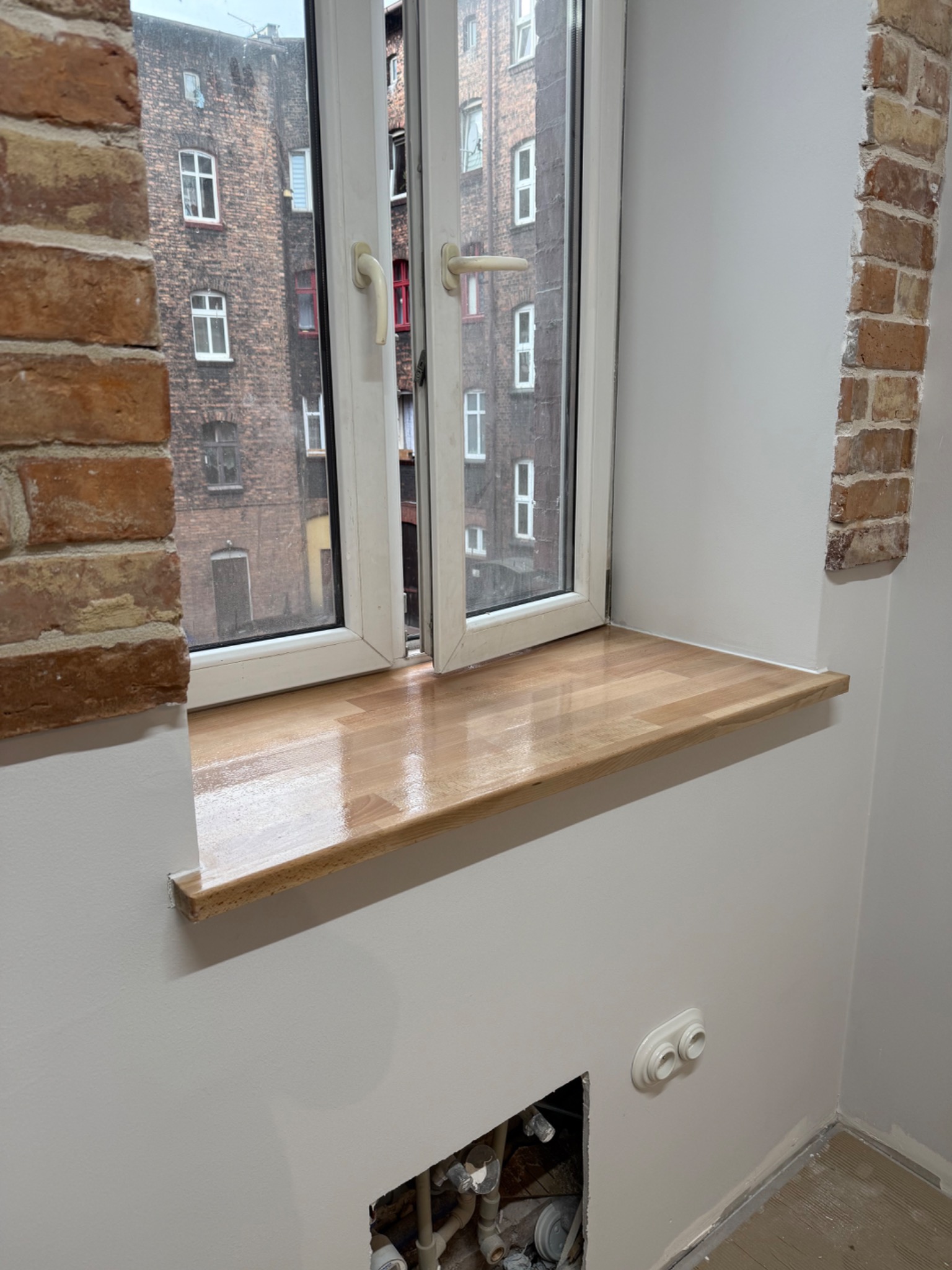 Nowy, drewniany parapet w oknie z widokiem na kamienicę, fragment ściany z cegły, gładź na ścianach, instalacja w ścianie przed wykończeniem.