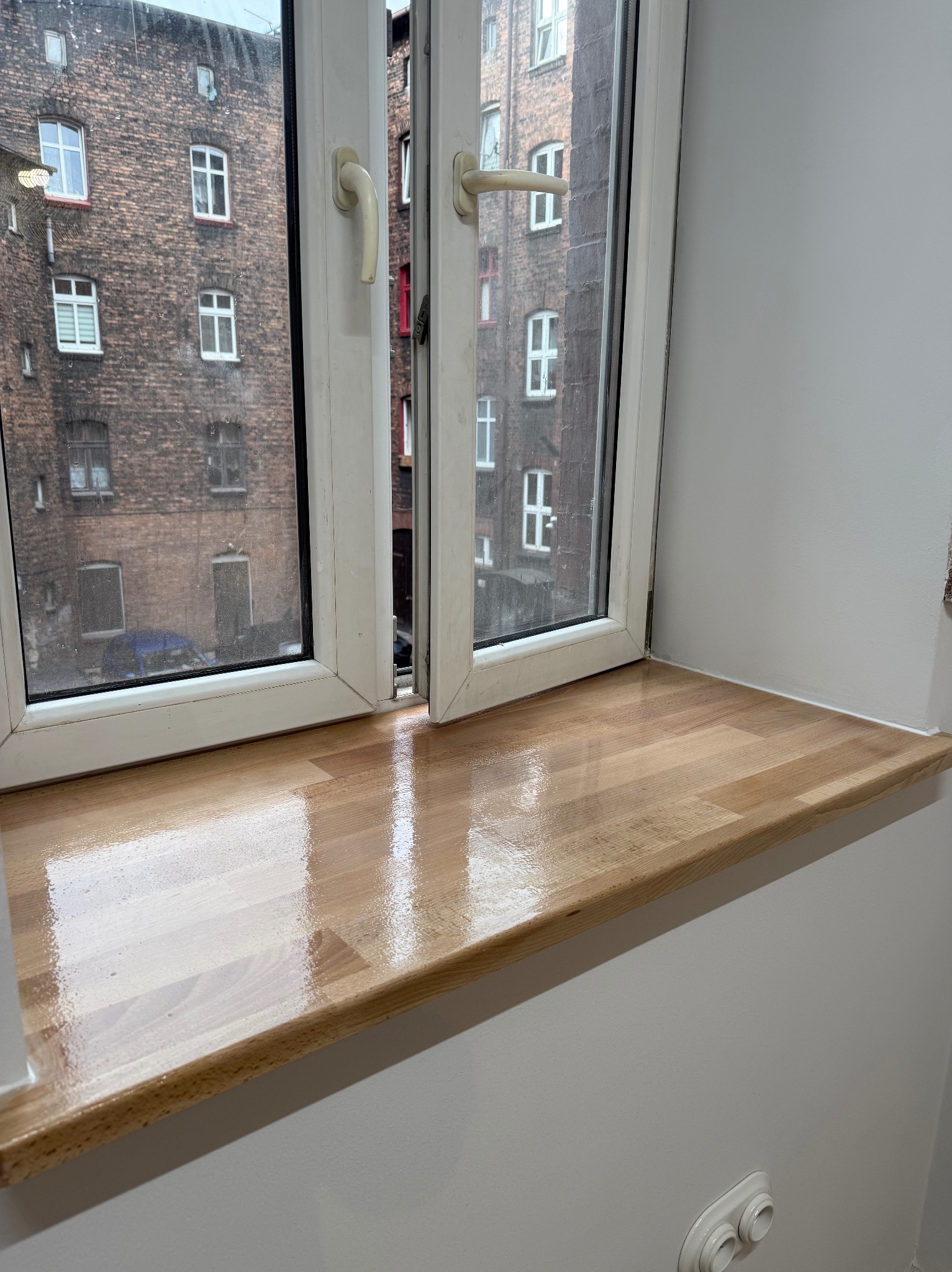 Jasny parapet z drewna klejonego, zamontowany pod białym oknem z widokiem na ceglany budynek. Świeżo pomalowana, gładka, biała ściana.