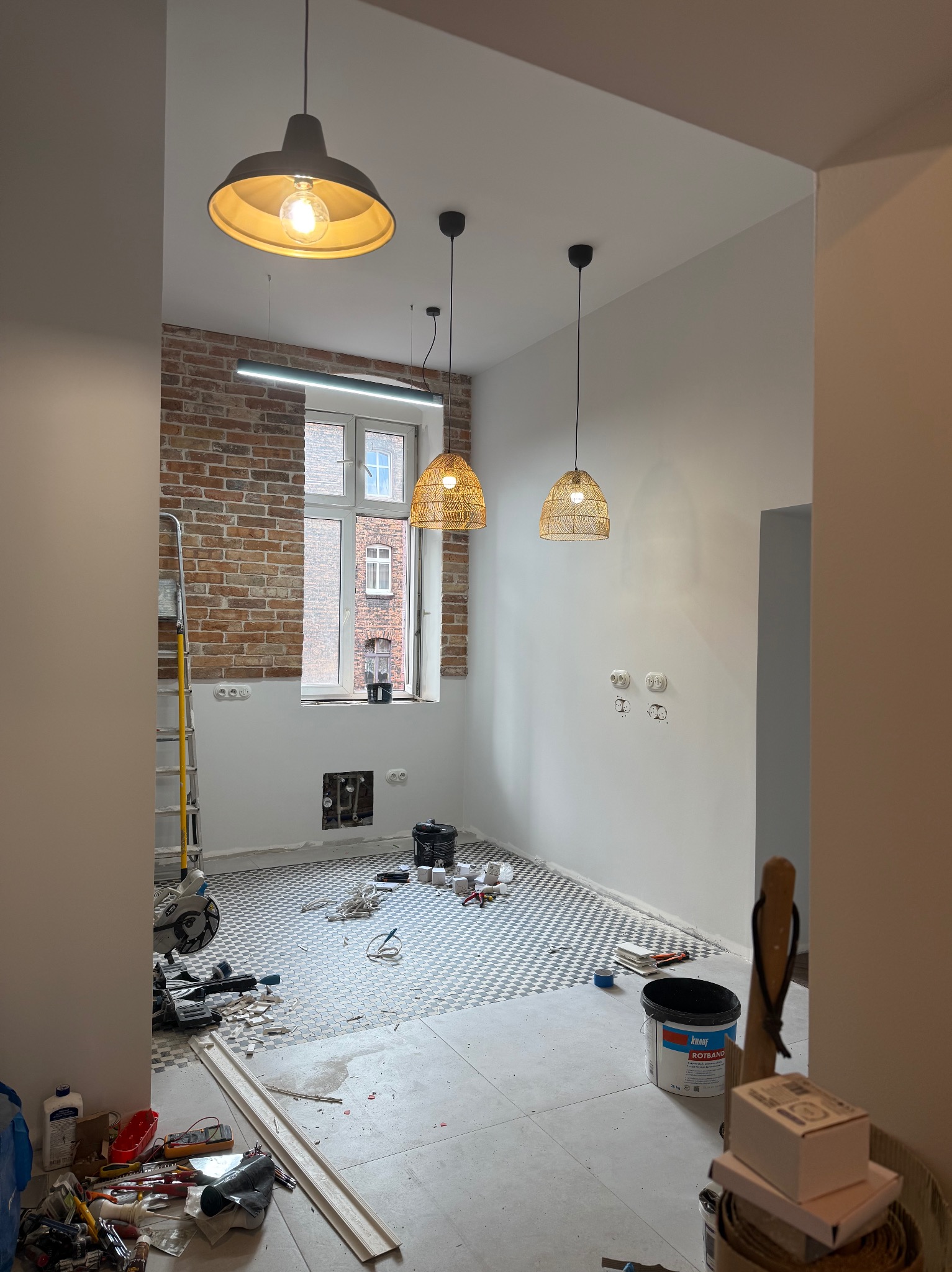 Remont wnętrza: ułożona mozaika podłogowa w pokoju z ceglaną ścianą, widoczne narzędzia i materiały budowlane, trzy lampy wiszące. Etap prac wykończeniowych.