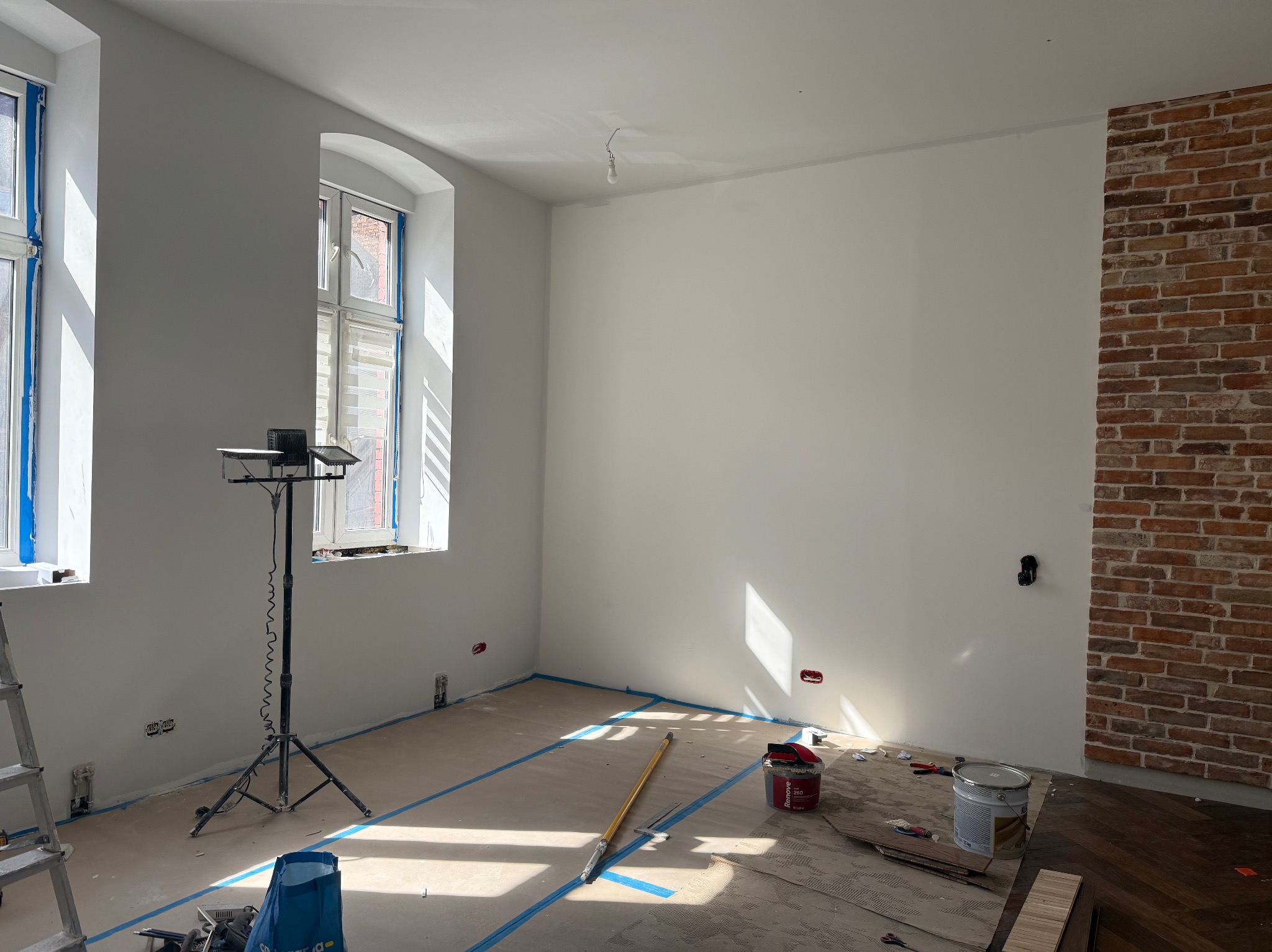 Pokój po malowaniu ścian na biało z widocznymi narzędziami malarskimi, zabezpieczonymi oknami i fragmentem ściany z cegły. Podłoga częściowo zabezpieczona folią.