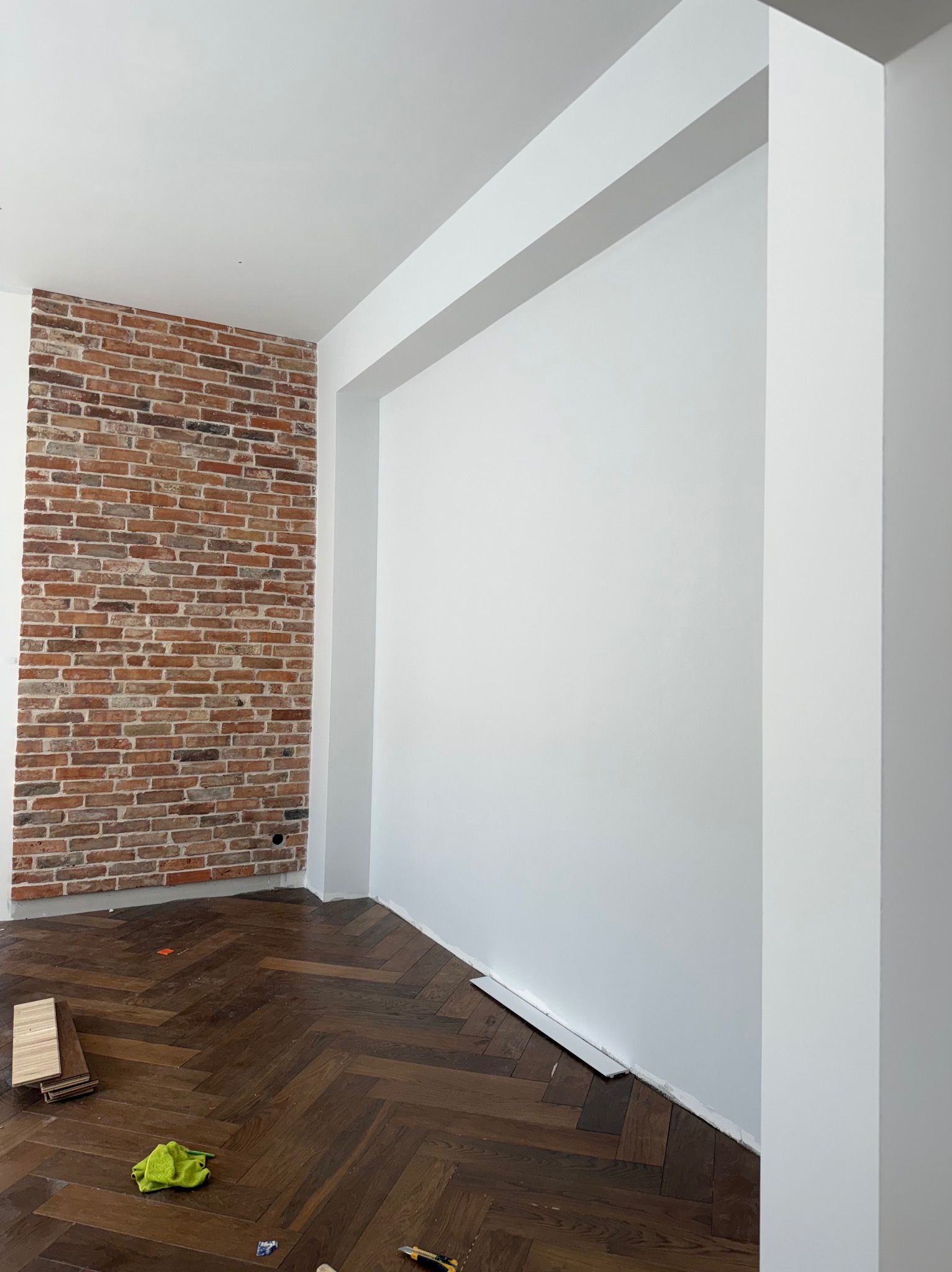 Pokój po remoncie z białą ścianą, fragmentem ściany z cegły i parkietem w jodełkę. Widoczne narzędzia i materiały wykończeniowe.