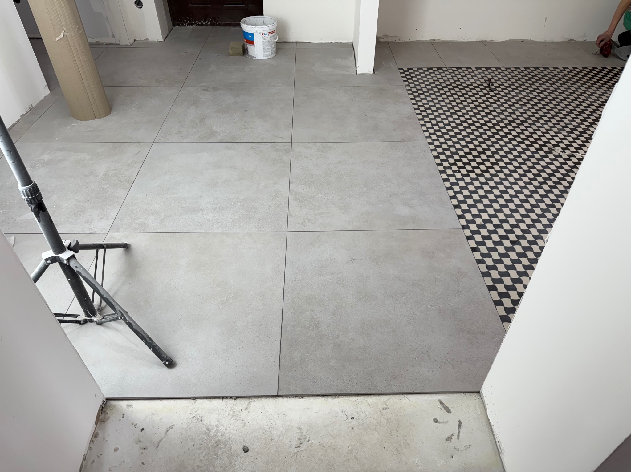 Ułożone szare płytki podłogowe w dużym formacie, kontrastujące z fragmentem podłogi w czarno-białą szachownicę. Widoczny statyw i wiadro z farbą.