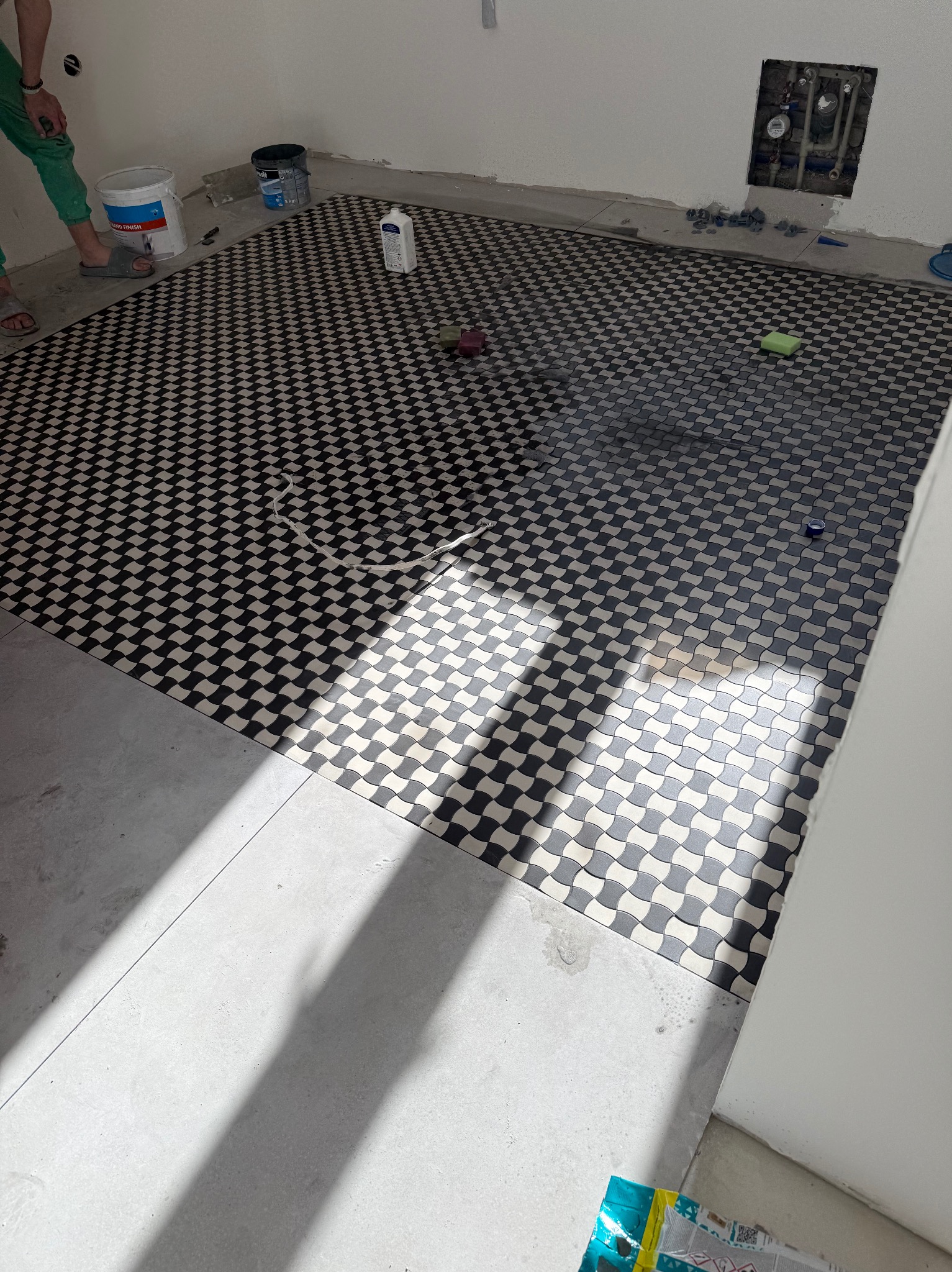 Ułożona podłoga z czarno-białej mozaiki w łazience, widoczne narzędzia glazurnicze i resztki materiałów budowlanych. Częściowo ułożone płytki podłogowe.