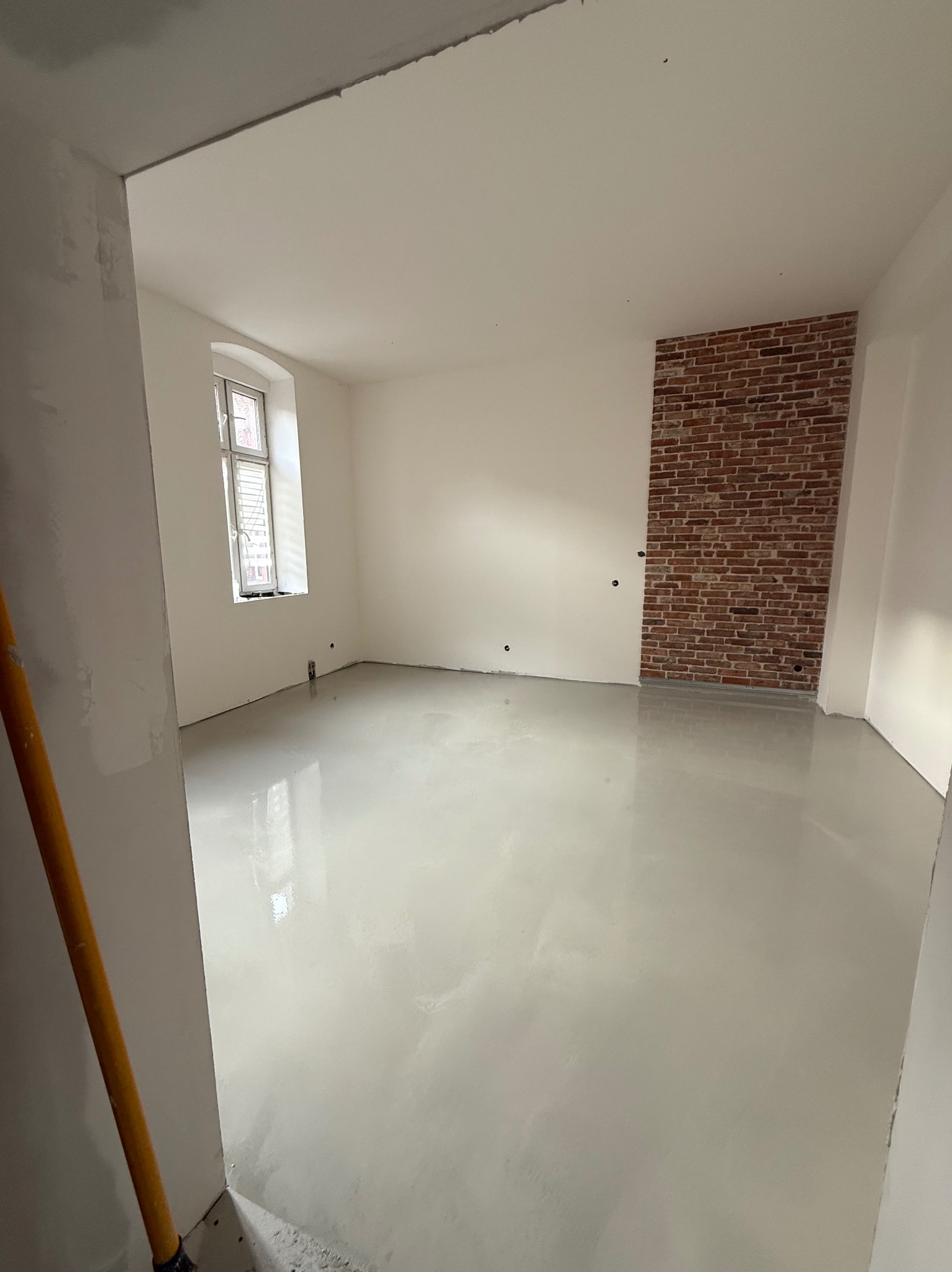 Białe, gładkie ściany i wylewka w pokoju z ceglaną ścianą akcentową. Okno z białymi ramami w tle. Minimalistyczny design wnętrza po remoncie.