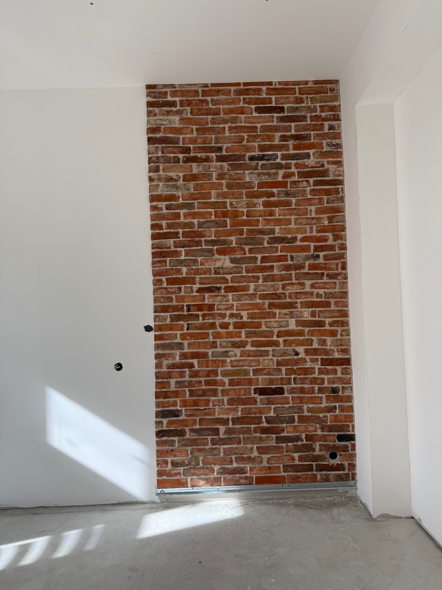 Ściana z cegły dekoracyjnej, częściowo wykończona, z widocznym profilem metalowym u dołu, na tle białej ściany i surowej podłogi. Otwory na gniazdka elektryczne.