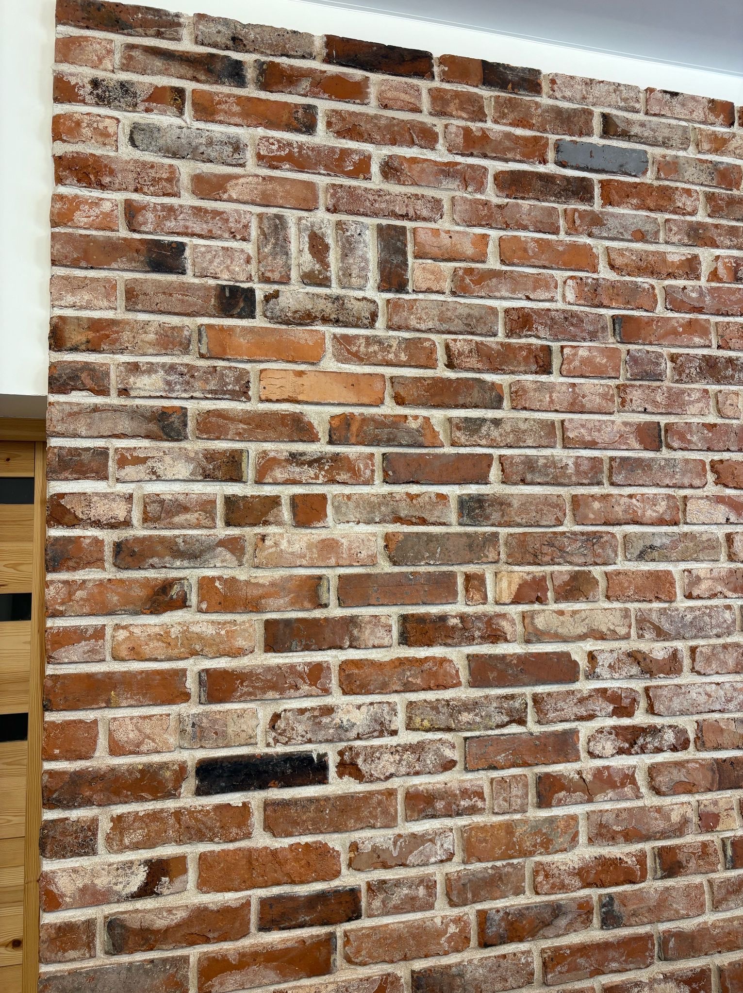 Ściana z czerwonej cegły z wyraźnymi spoinami, fragment drewnianej ramy drzwi po lewej stronie. Dekoracyjne wykończenie wnętrza z naturalnych materiałów.