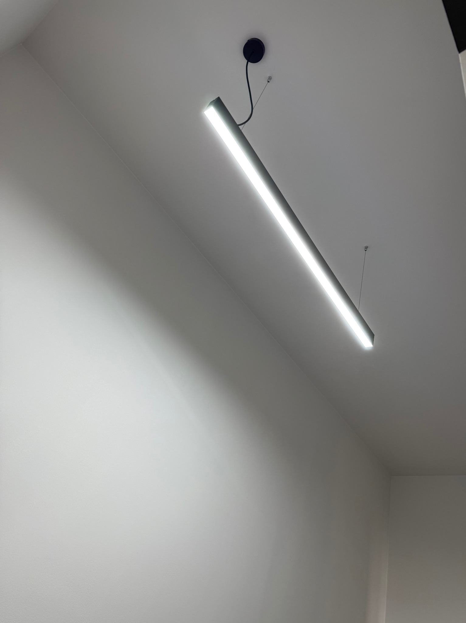 Minimalistyczne wnętrze z oświetleniem LED: Długa lampa wisząca, równomiernie oświetlająca białą ścianę i sufit. Nowoczesny design, stonowana kolorystyka.