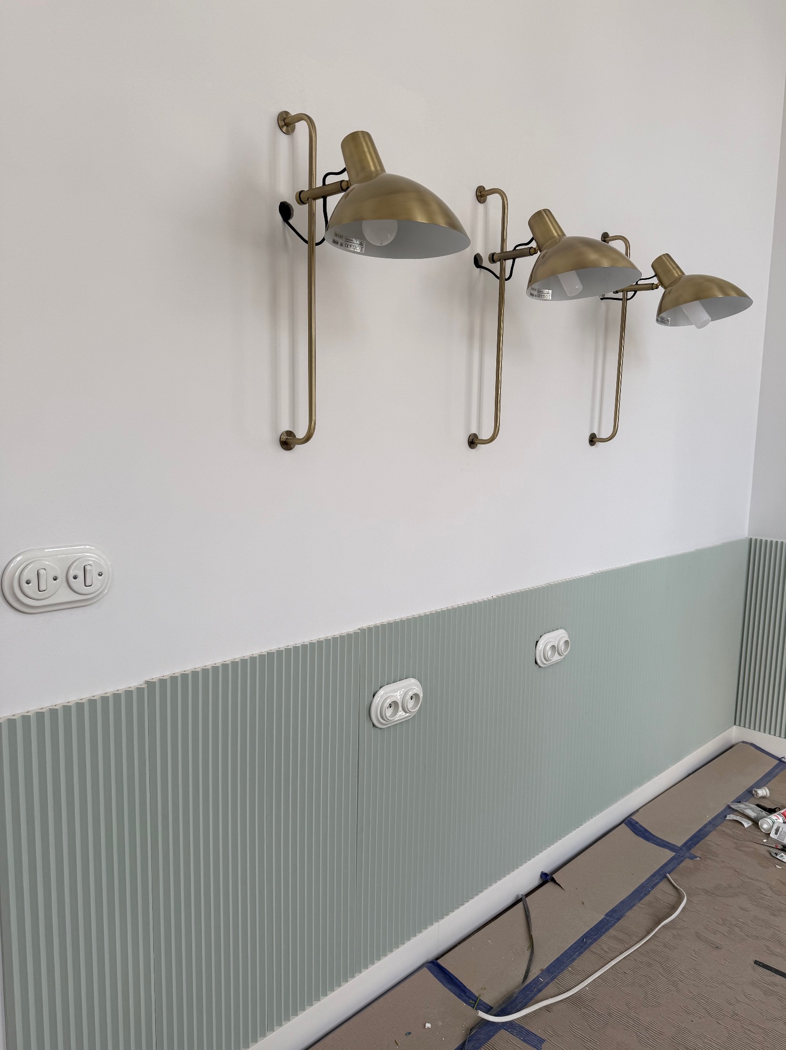 Ściana z dekoracyjną, pionowo żłobioną boazerią w odcieniach szarości, z trzema złotymi lampami ściennymi i białymi gniazdkami. Widoczne zabezpieczenia podłogi.