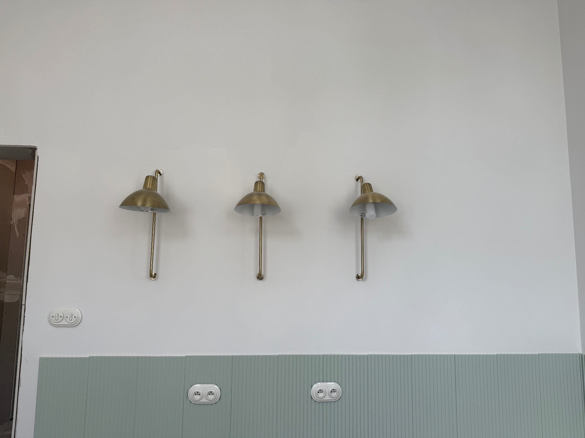 Wykończona ściana z trzema złotymi lampami ściennymi, listwami dekoracyjnymi w kolorze miętowym i białymi gniazdkami elektrycznymi. Widoczny fragment niezakończonego remontu.