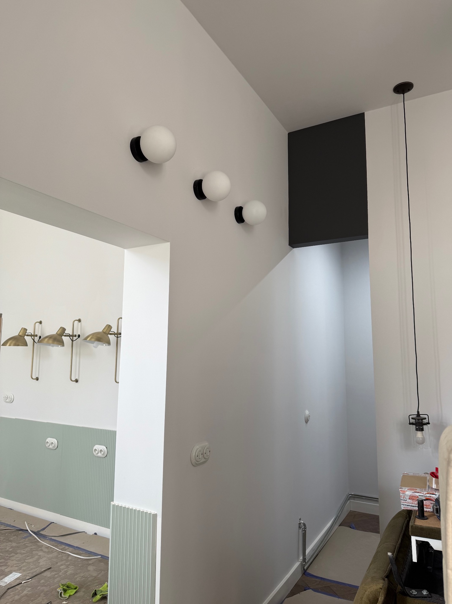Wnętrze w trakcie wykańczania: białe ściany z dekoracyjnymi lampami kulami i złotymi kinkietami. Widoczne gniazdka, listwy przypodłogowe i fragment podłogi z zabezpieczeniami.