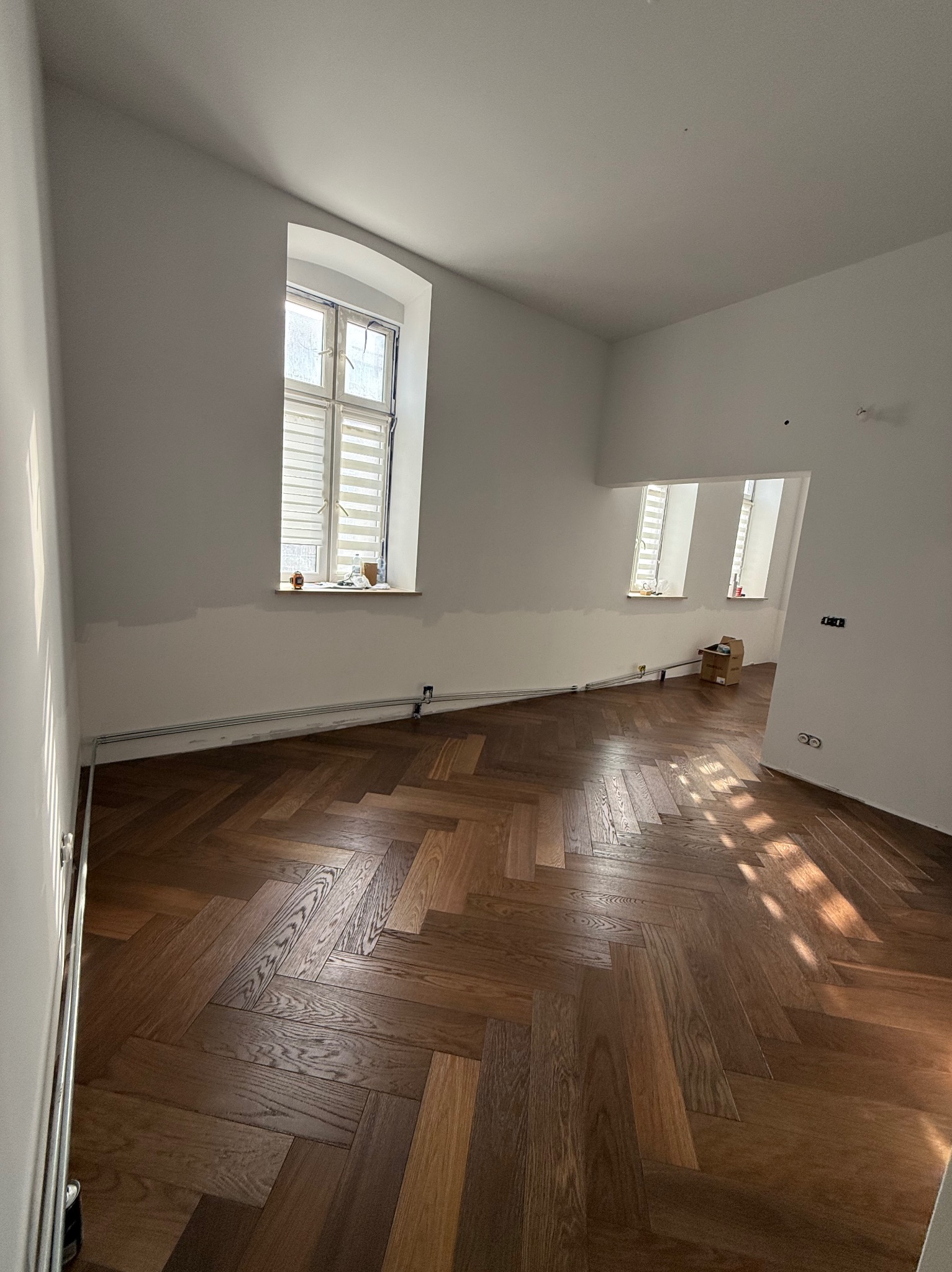 Parkiet w jodełkę w jasnym pokoju z wysokimi oknami. Ściany przygotowane do malowania, widoczne ślady po taśmie malarskiej. Częściowo wykończone wnętrze.