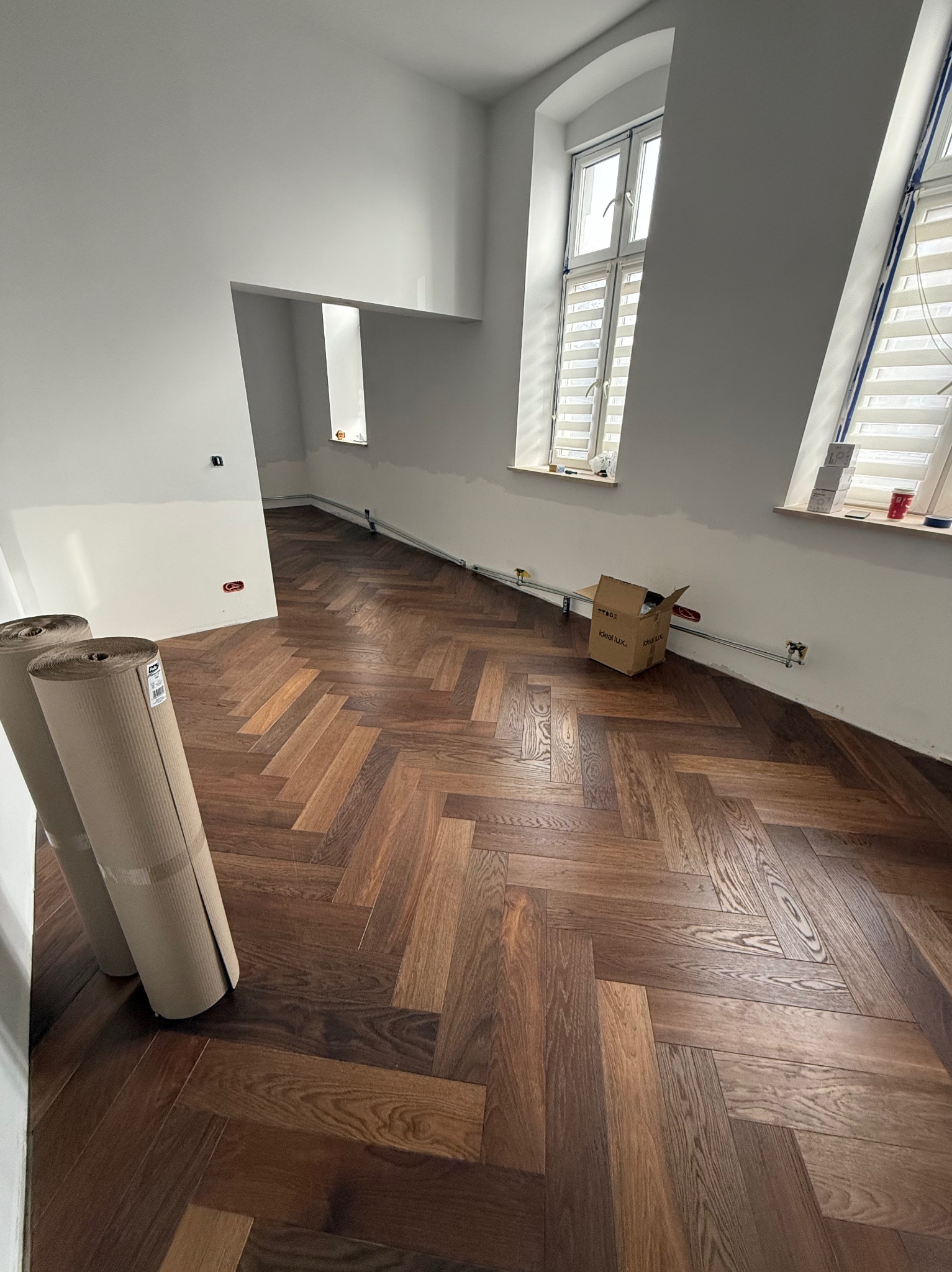 Ukończona podłoga z parkietu w jodełkę w jasnym wnętrzu. Widoczne rolki z materiałem zabezpieczającym i karton. Dwa okna z żaluzjami w tle.
