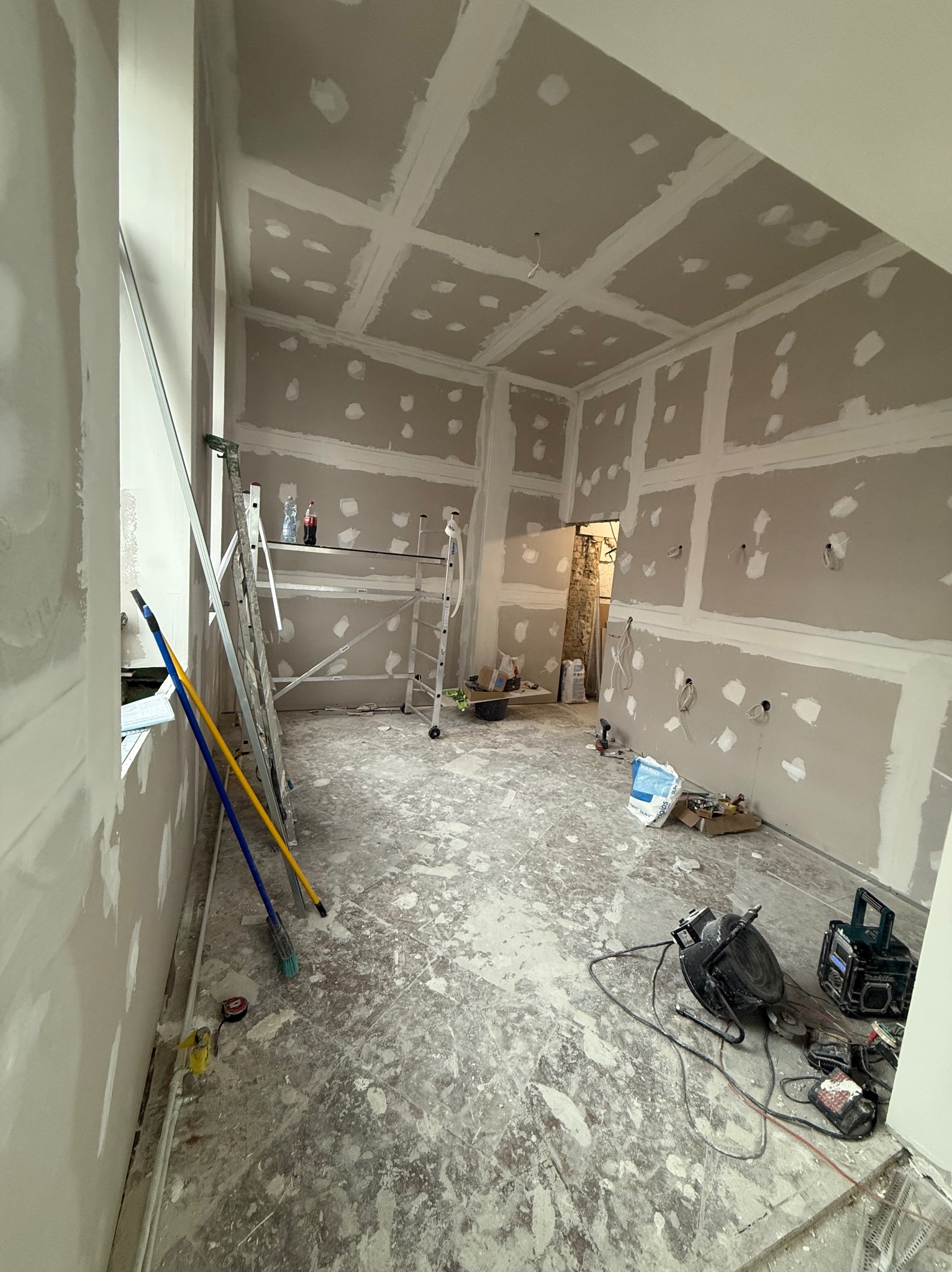 Pokój w trakcie prac z płytami gipsowo-kartonowymi na ścianach i suficie. Widoczne narzędzia, drabina i rusztowanie. Podłoga pokryta resztkami materiałów budowlanych.