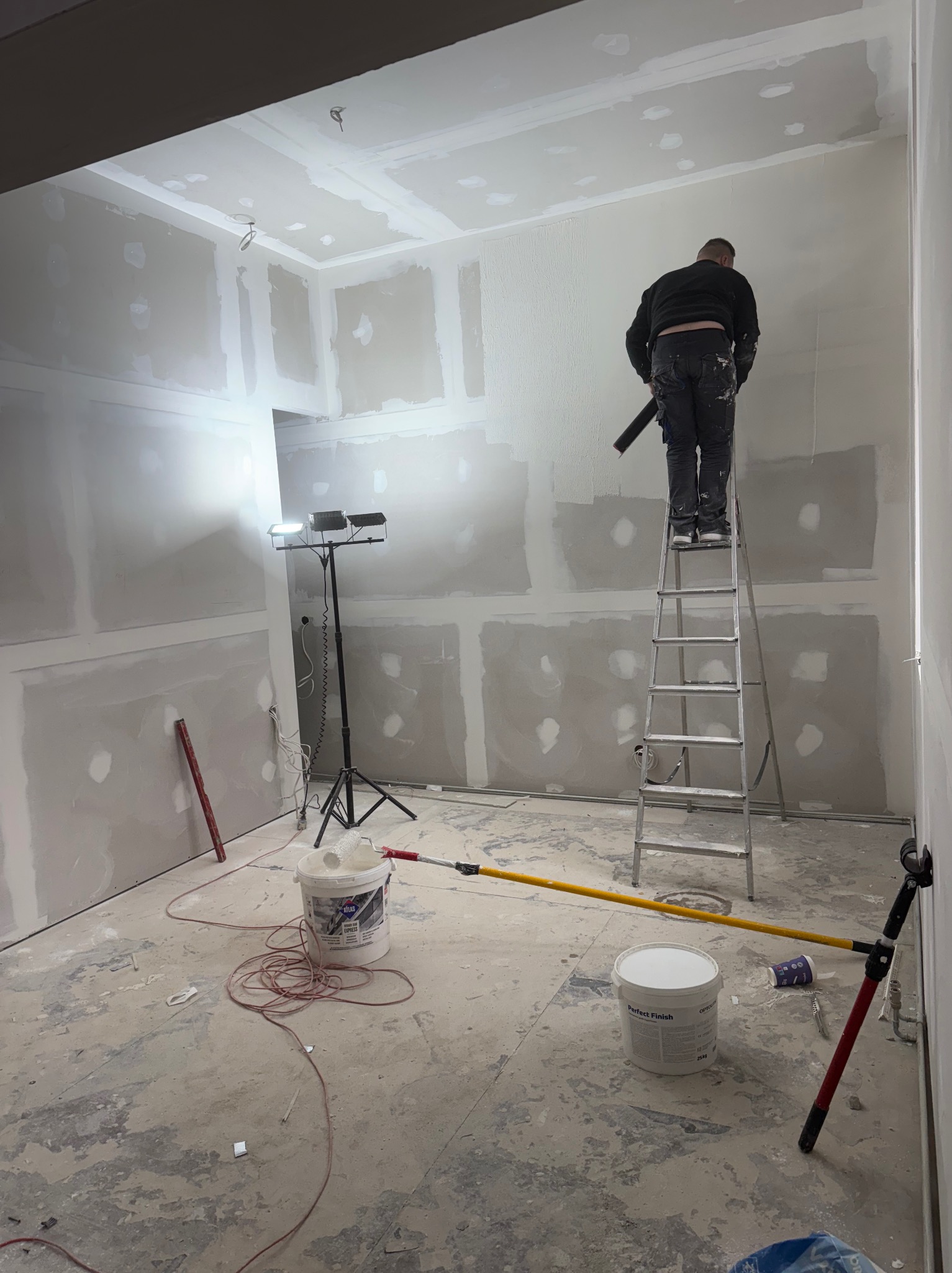 Prace wykończeniowe w nowym mieszkaniu: mężczyzna na drabinie szpachluje ścianę z płyt gipsowo-kartonowych, widoczne narzędzia, oświetlenie i wiaderka z masą szpachlową.