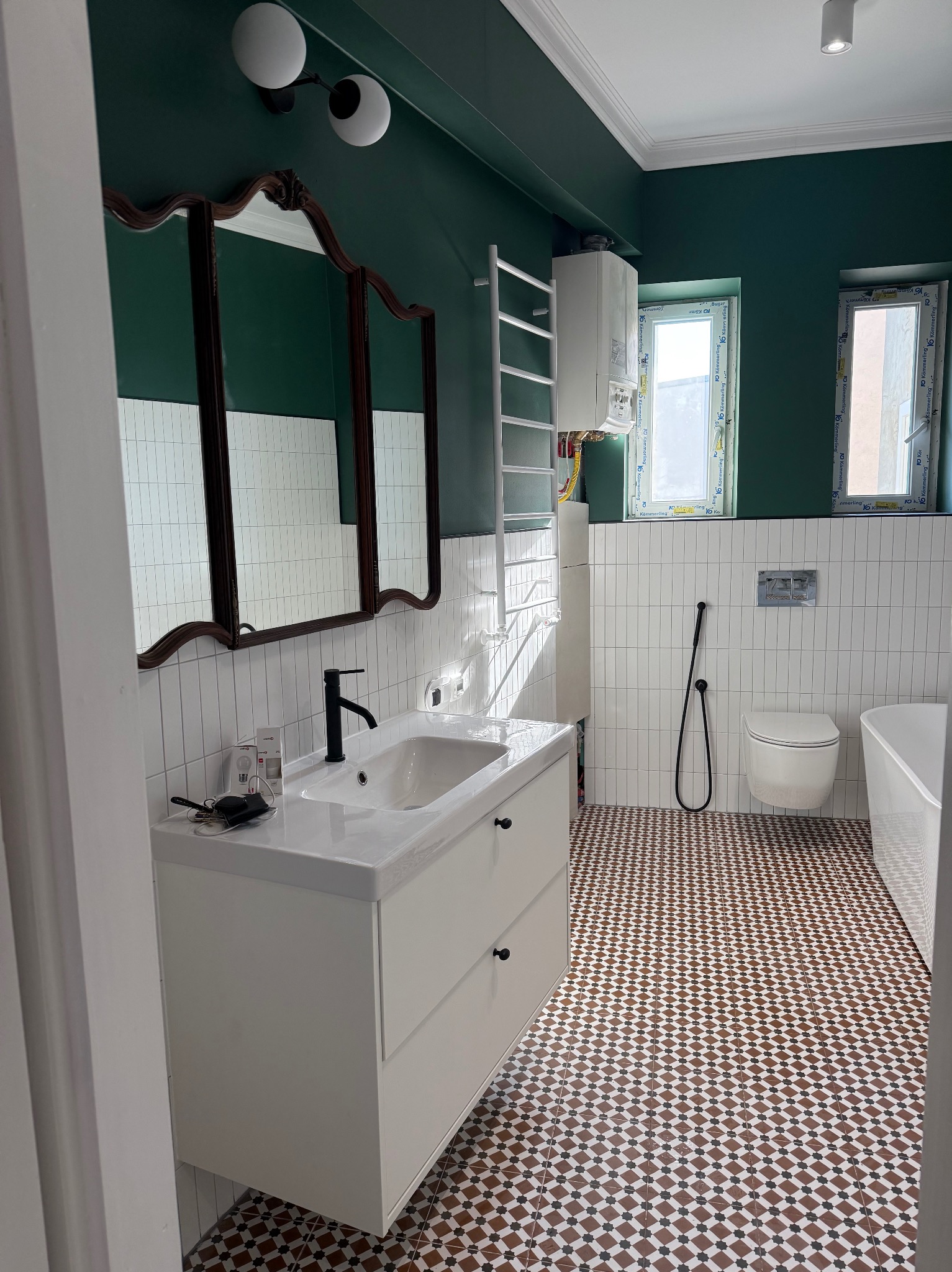 Stylowa łazienka po remoncie: białe płytki pionowe, dekoracyjna podłoga, ciemnozielone ściany, ozdobne lustro i nowoczesna armatura. Uwagę przyciąga nietypowy wzór podłogi.