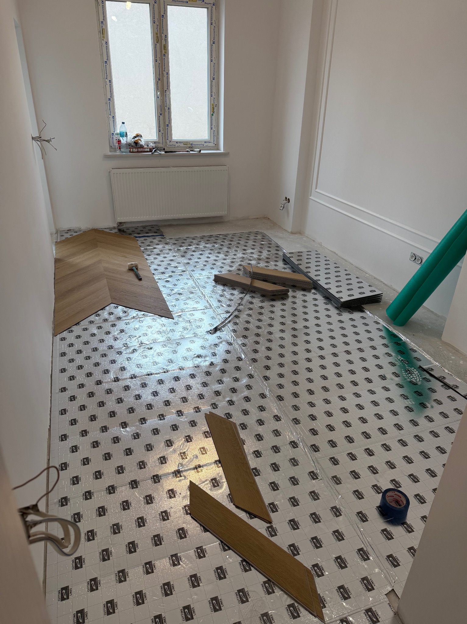 Układanie drewnianej podłogi w jodełkę na folii izolacyjnej. Widoczne narzędzia i fragmenty paneli. W tle okno z białymi ramami i grzejnik.