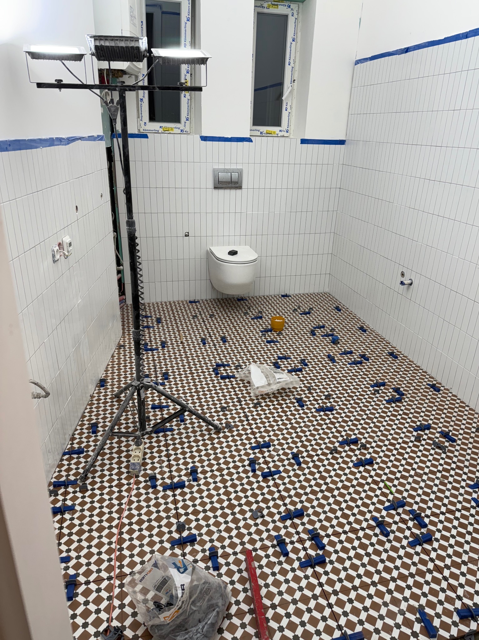 Układanie mozaiki w łazience w Chorzowie. Na podłodze brązowo-biała szachownica z niebieskimi klinami poziomującymi. Ściany wyłożone białymi, pionowymi płytkami.