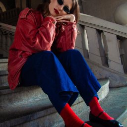 Pinky Foto - Amelia Jaworska - Portret kobiety w czerwonej skórzanej kurtce i okularach, siedzącej na betonowych schodach z balustradą, w intensywnym oświetleniu.