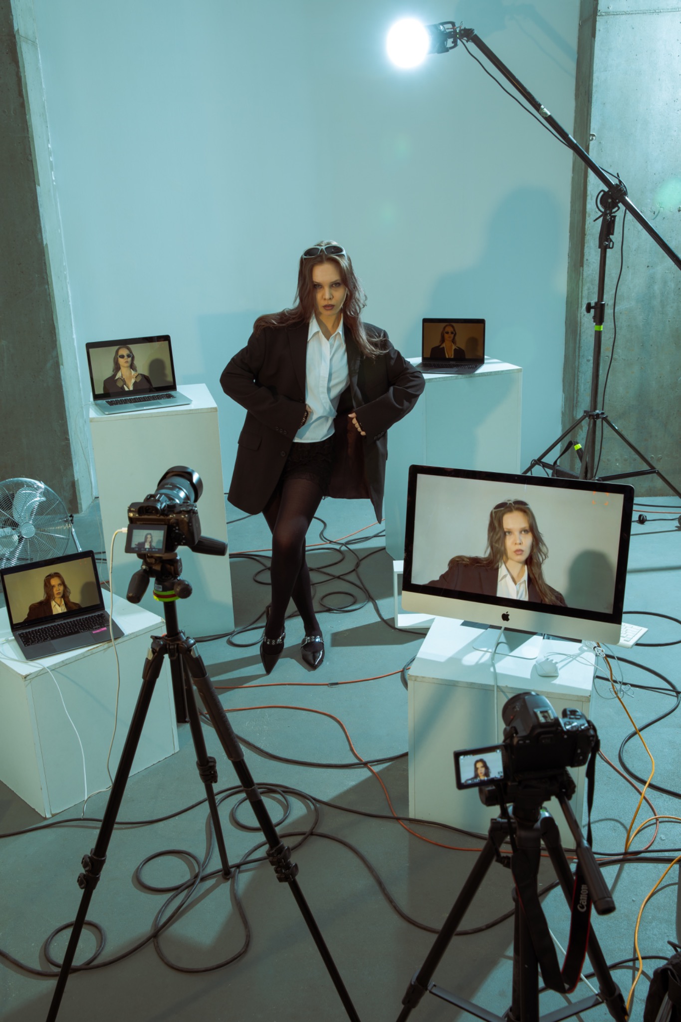 Za kulisami sesji zdjęciowej: modelka w oversize'owej marynarce pozuje otoczona monitorami i aparatami na statywach, oświetlona lampą studyjną. Widoczne kable i wentylator.