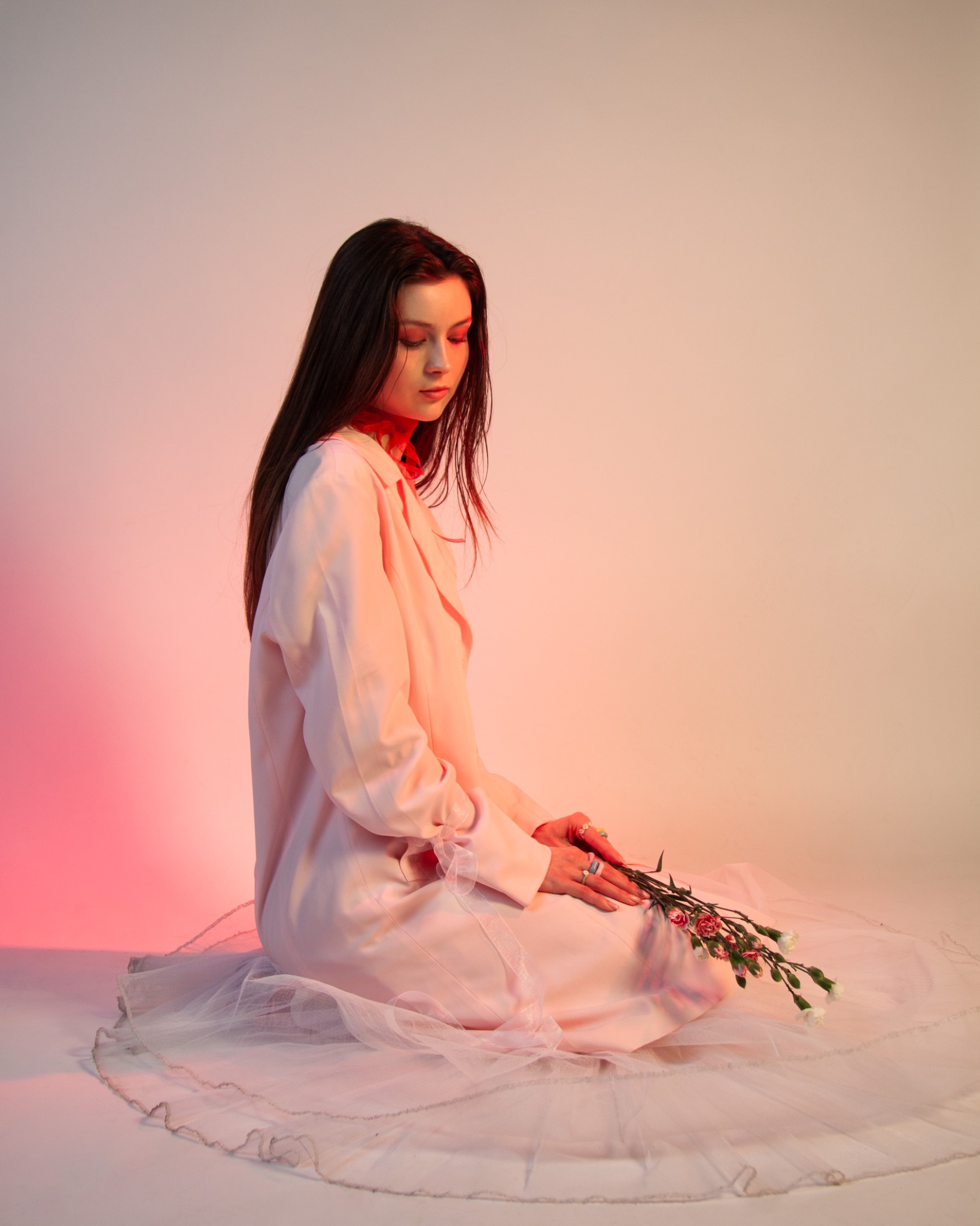 Kobieta w pastelowej stylizacji siedzi na tiulowej tkaninie, trzymając gałązkę kwiatów na tle gradientowego oświetlenia w odcieniach różu i beżu.