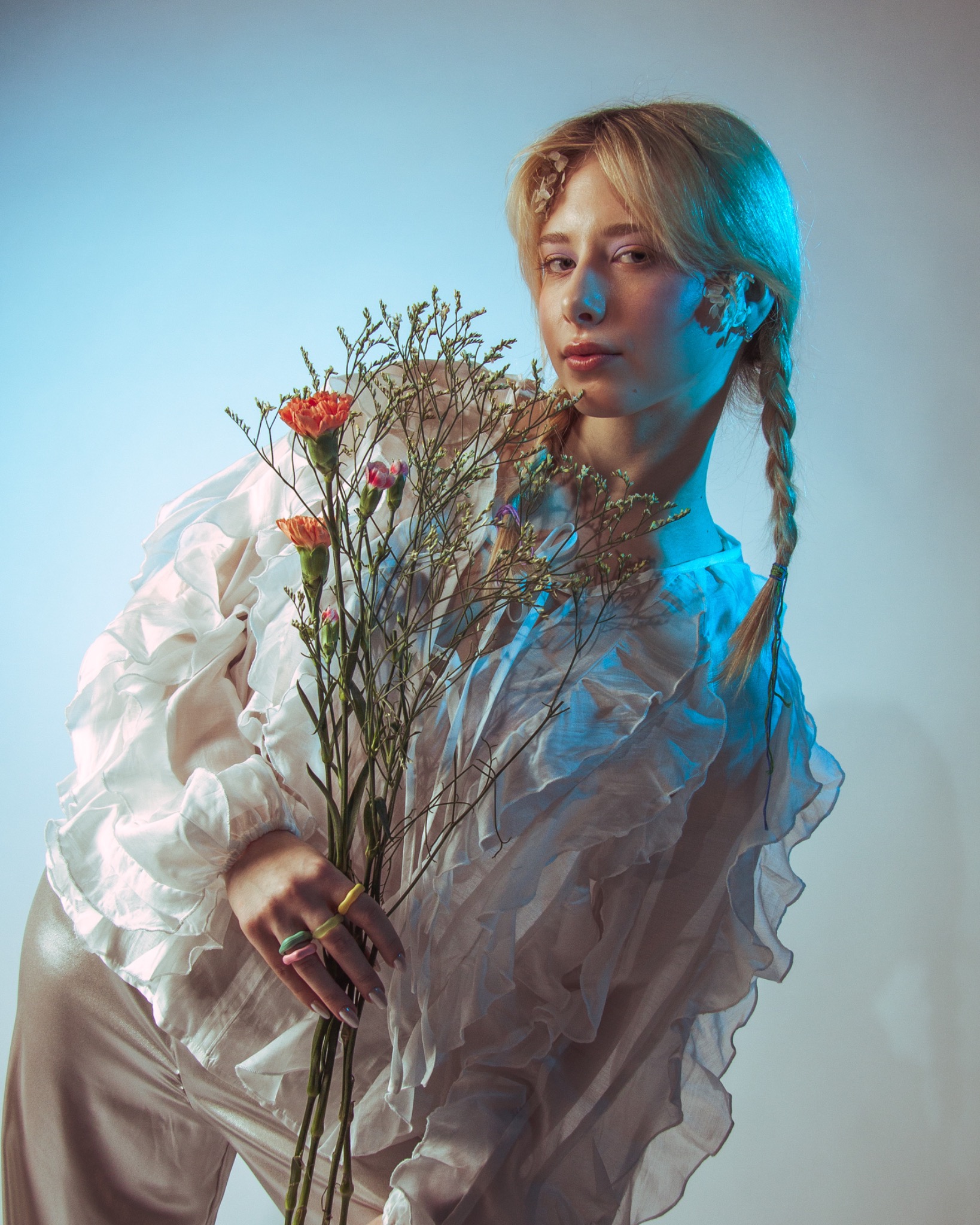 Portret kobiety z blond warkoczami, trzymającej bukiet kwiatów w pastelowych kolorach, oświetlonej niebieskim światłem i ubranej w białą bluzkę z falbanami.