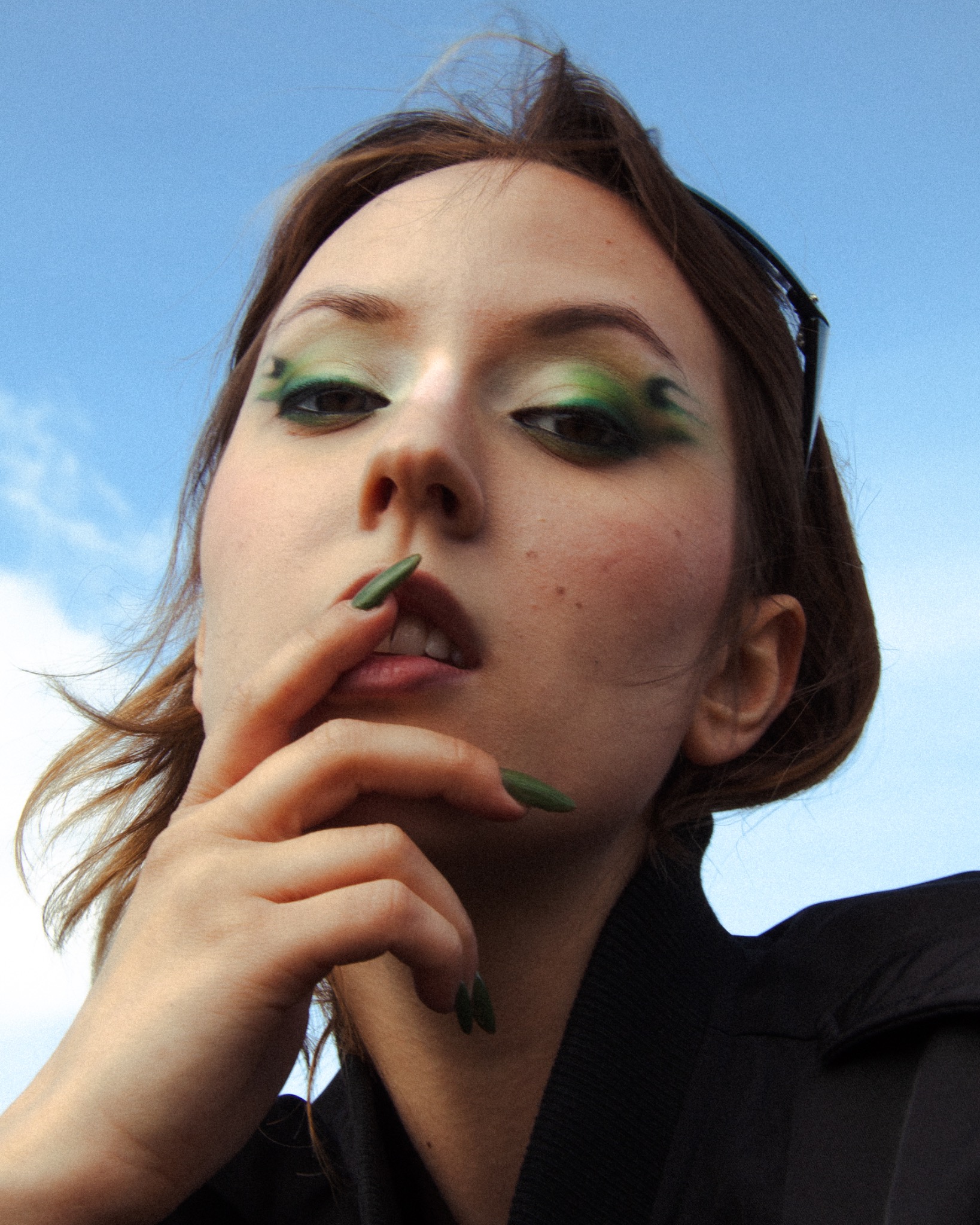 Portret kobiety z zielonym makijażem i paznokciami na tle błękitnego nieba w Mielcu. Ujęcie z dołu podkreśla artystyczny styl i nowoczesny wygląd.
