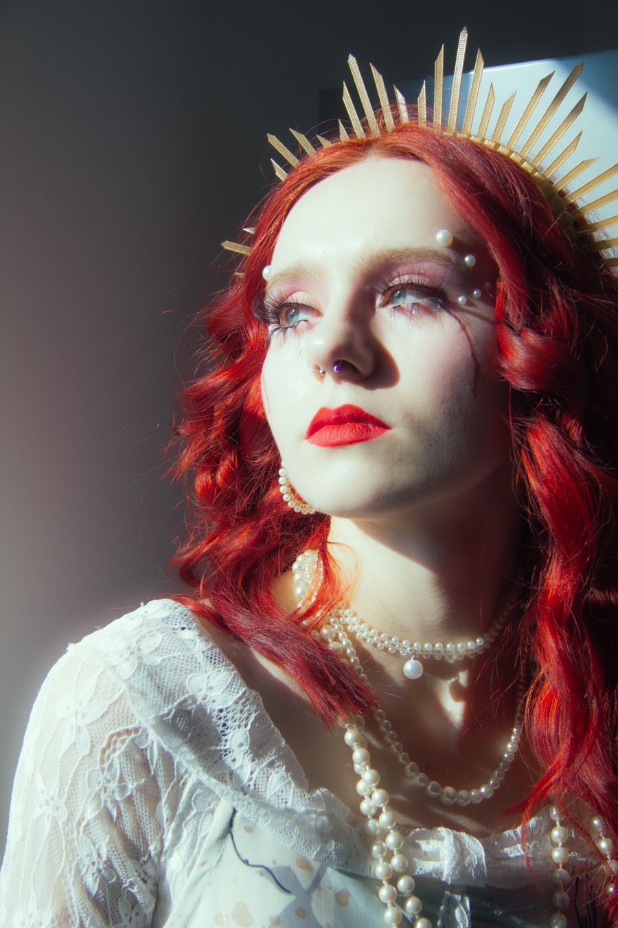 Portret kobiety z rudymi włosami, w koronie i perłach, z artystycznym makijażem, w oświetleniu studyjnym, spoglądającej w górę w lewo, z kolczykiem w nosie.