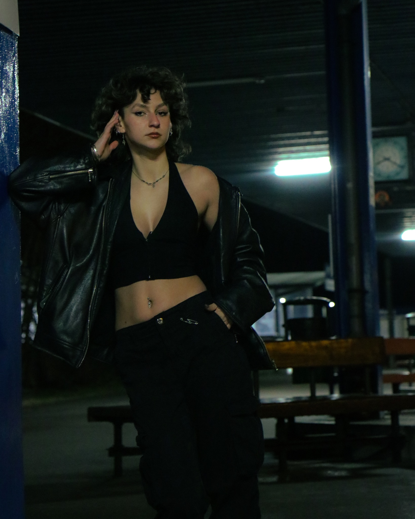 Portret kobiety w czarnym stroju i skórzanej kurtce, opierającej się o niebieski filar na tle dworca. Mroczny, miejski klimat, oświetlenie nocne, wyraźny makijaż.