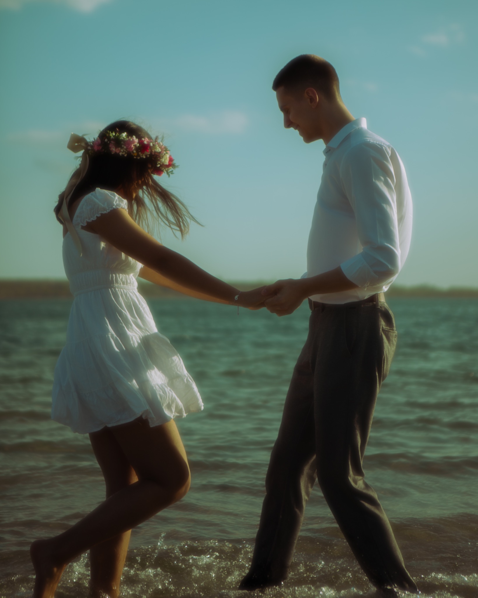 Para trzymająca się za ręce spaceruje po płytkiej wodzie. Kobieta ma wianek z kwiatów. Delikatne, naturalne światło podkreśla romantyczny nastrój.