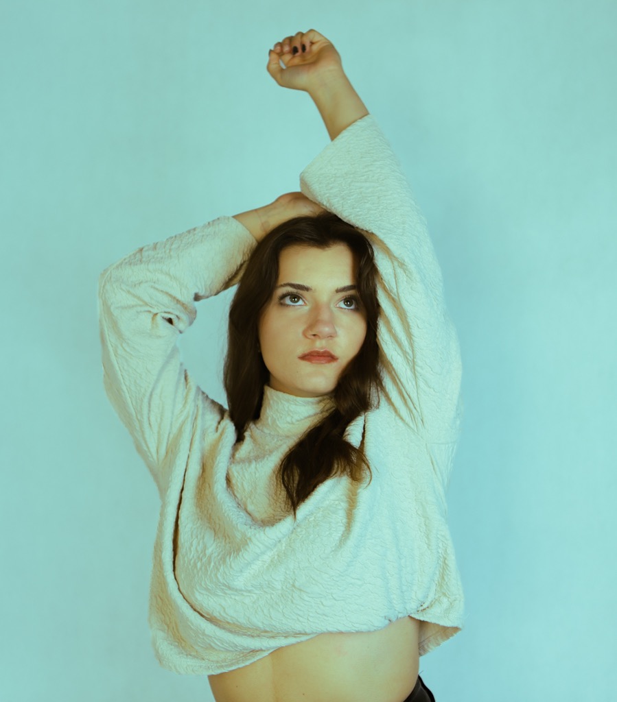 Portret kobiety w beżowej bluzce z rękami uniesionymi nad głową na tle błękitnej ściany. Ujęcie od dołu, podkreślające wyraz twarzy i stylizację.