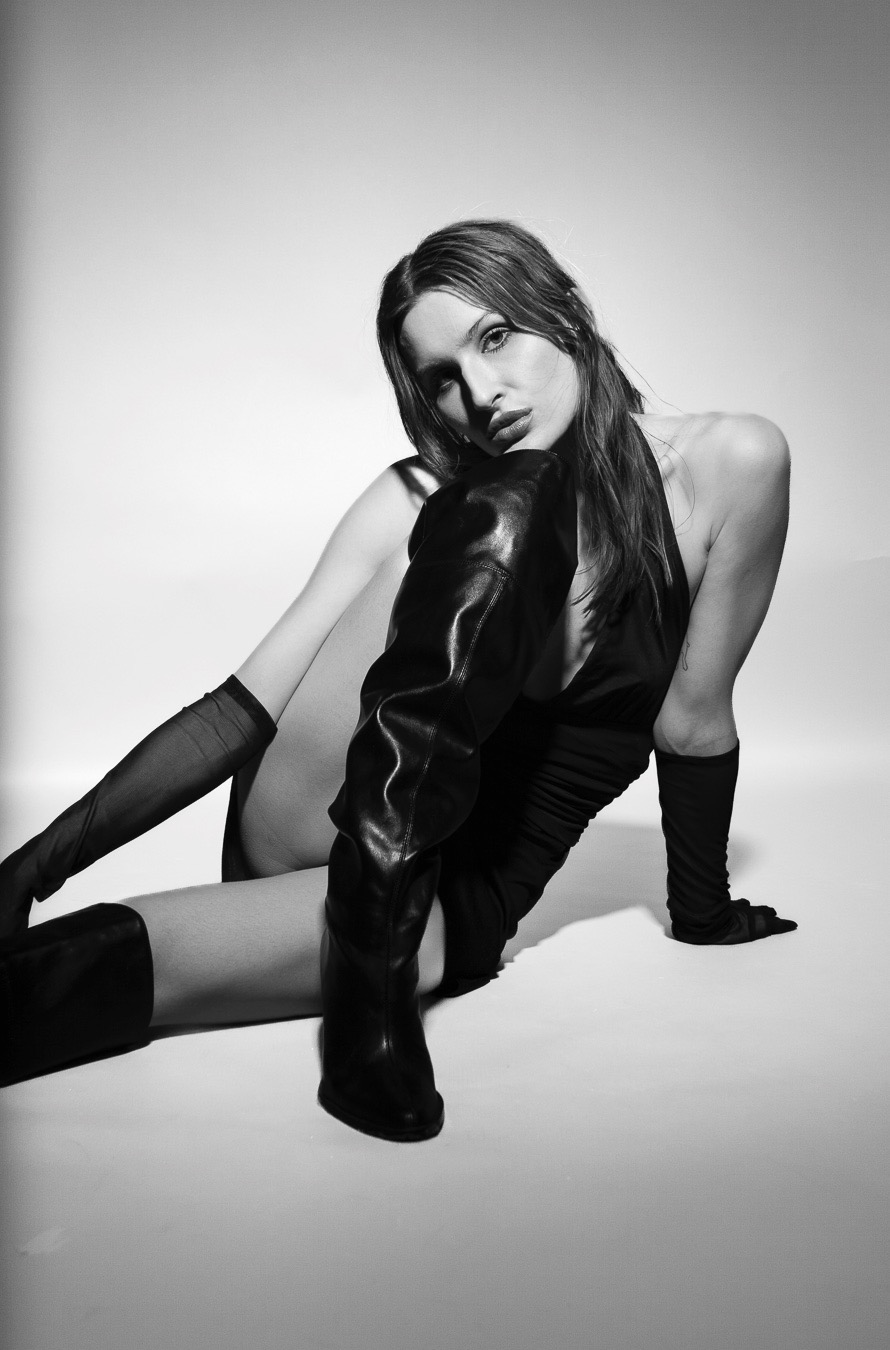 Monochromatyczne zdjęcie kobiety w czarnym body i wysokich skórzanych butach, pozującej na białym tle w studyjnym oświetleniu. Wyrazisty makijaż i pewna poza.