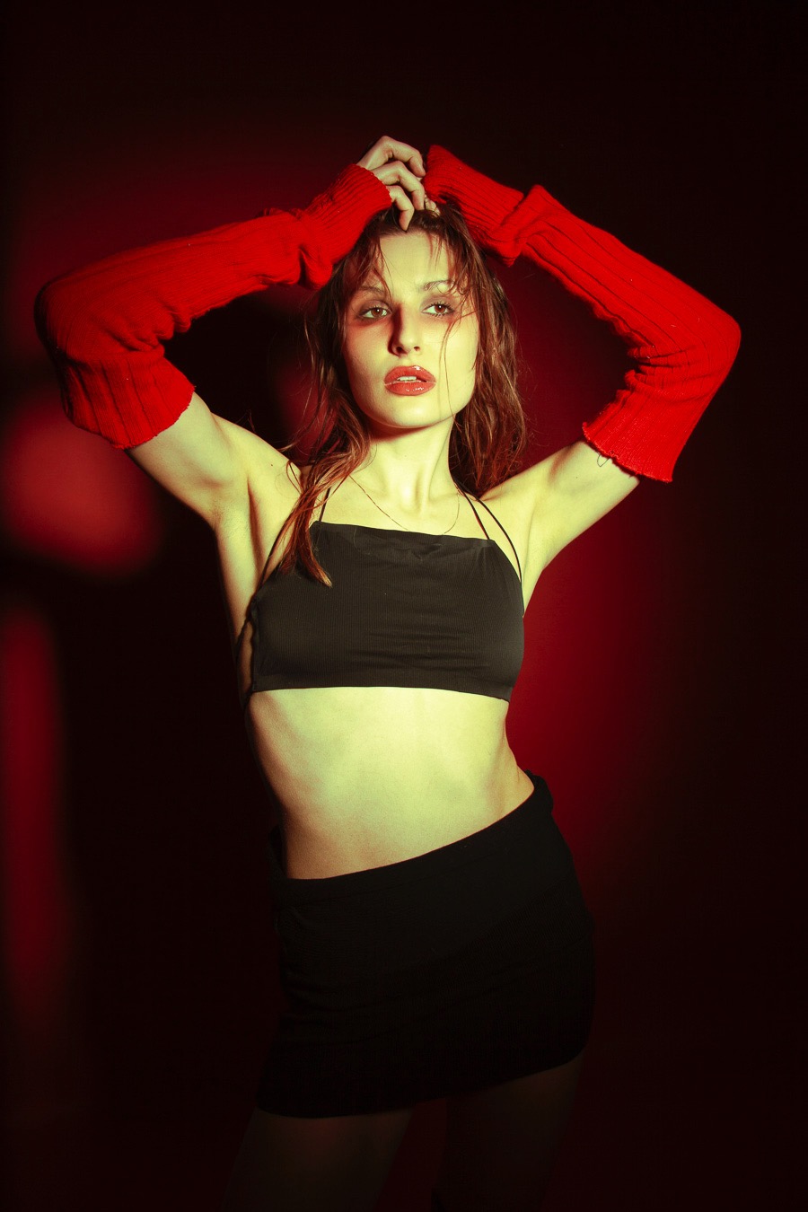 Portret kobiety w czerwonych rękawiczkach na tle ciemnego studia w Mielcu. Modelka ma mokre włosy i pozuje z rękami uniesionymi nad głową. Czerwone oświetlenie.