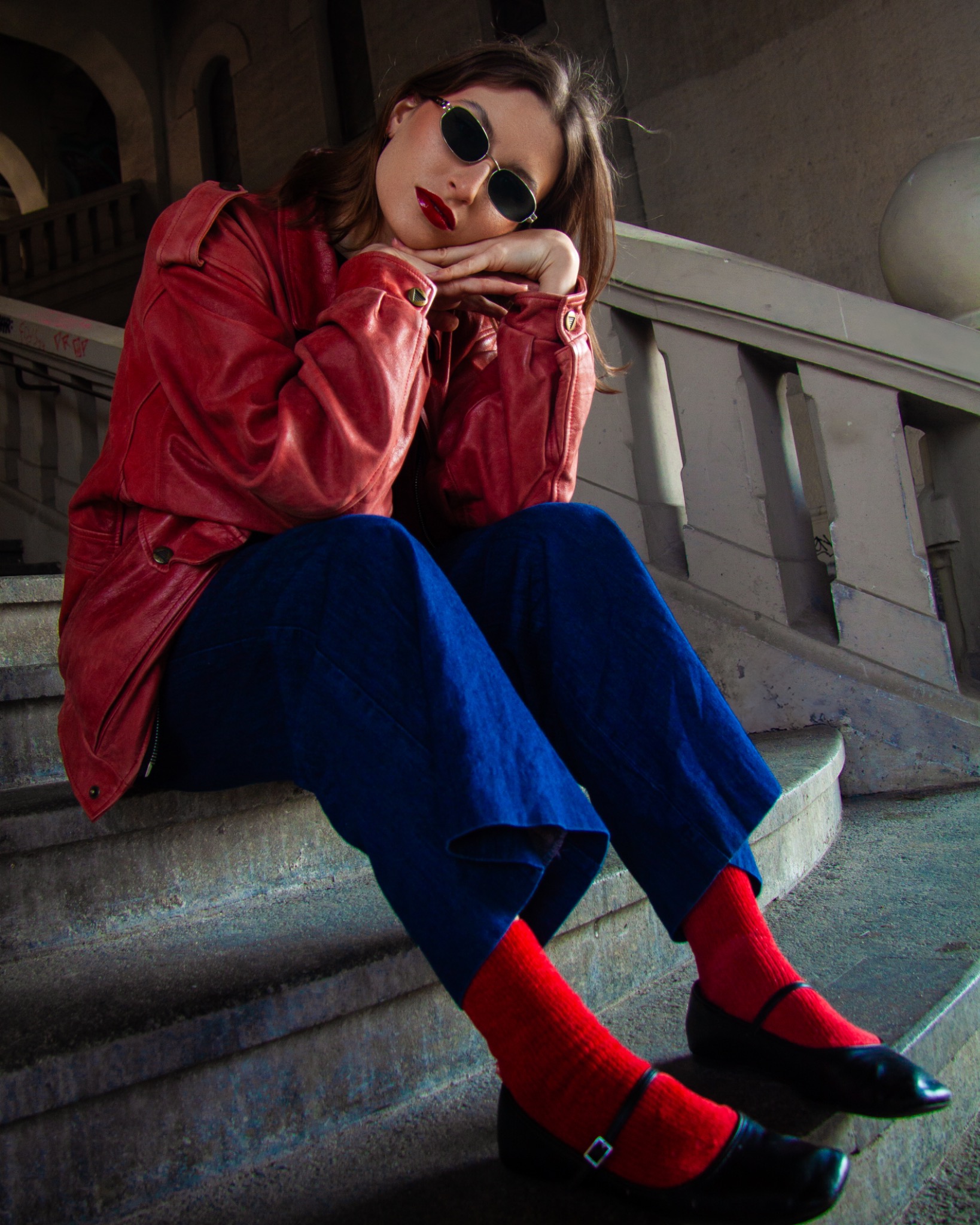 Portret kobiety w czerwonej skórzanej kurtce i okularach, siedzącej na betonowych schodach z balustradą, w intensywnym oświetleniu.