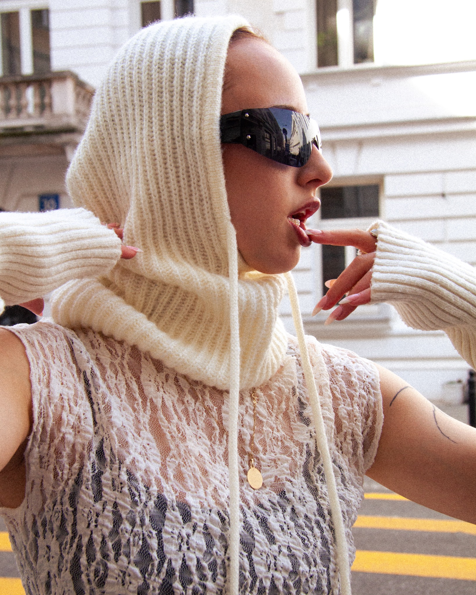 Kobieta w kremowym dzierganym kominie z kapturem i futurystycznych okularach, w przezroczystej bluzce, na tle miejskiej architektury.