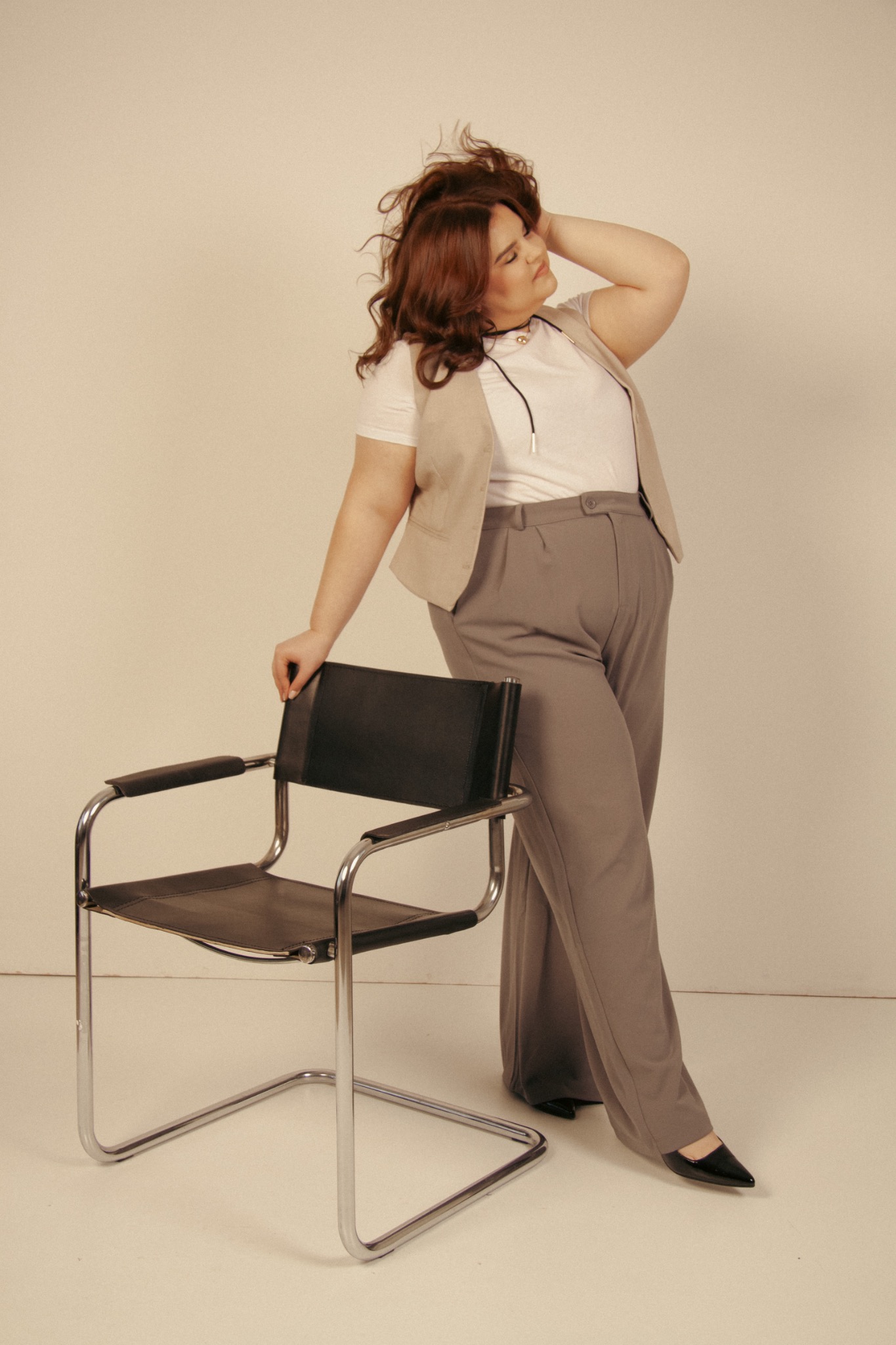 Kobieta plus size w stylizacji biznesowej pozuje opierając się o krzesło. Uśmiech, rozpuszczone włosy i stonowana kolorystyka tworzą profesjonalny, modny wizerunek.