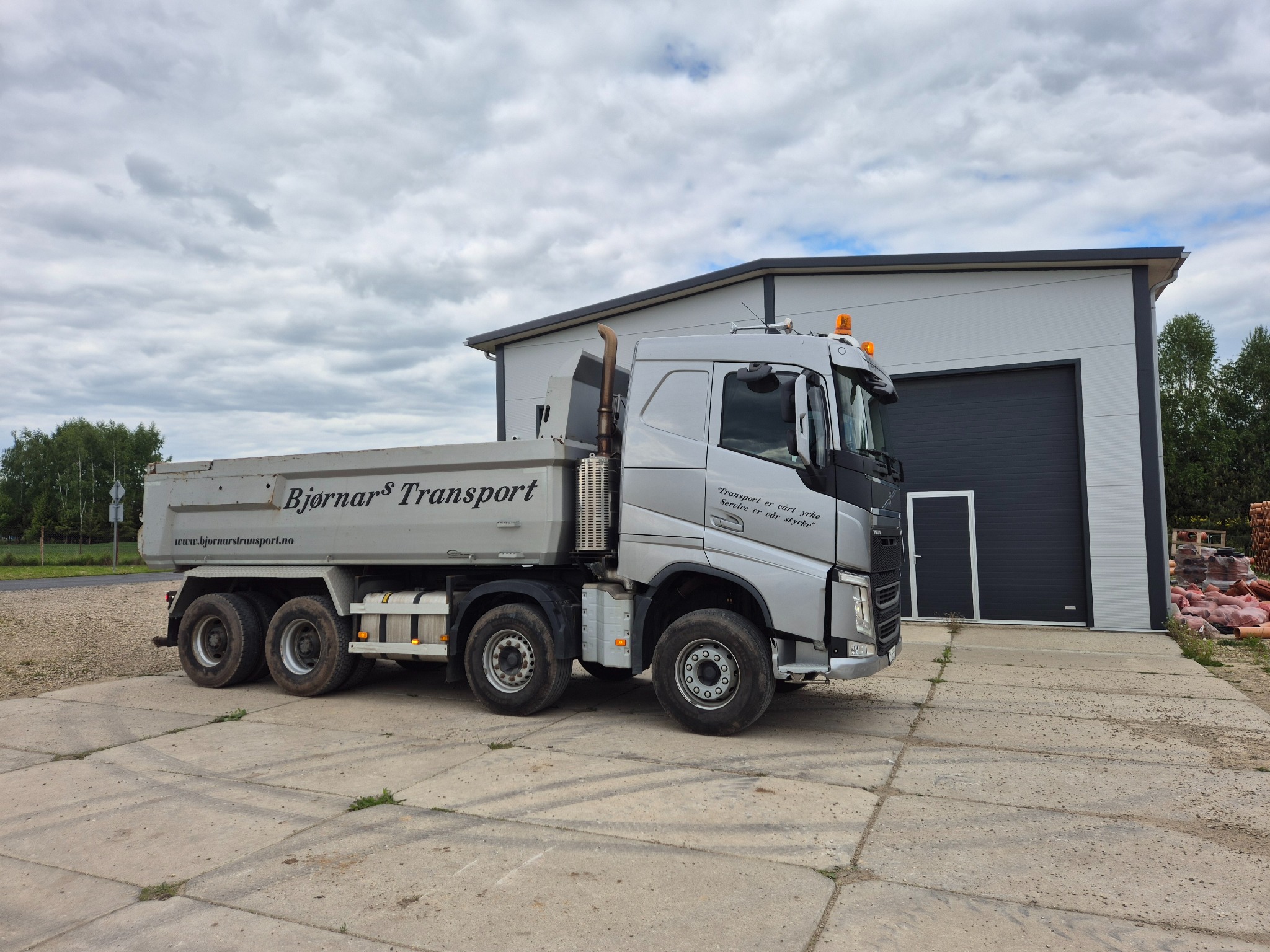 Srebrna wywrotka marki Volvo z napisem Bjornars Transport stoi na betonowym placu przed budynkiem z szarą bramą. Widoczne opony i błotniki.