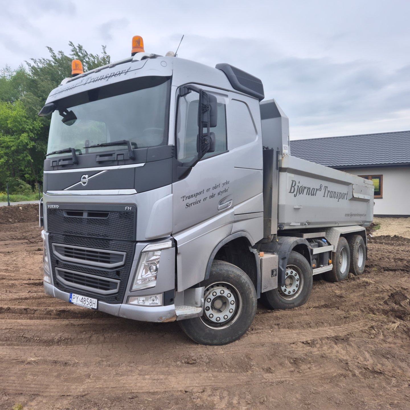 Szary, czysty wywrotka Volvo FH na placu budowy, z napisem 'Bjørnars Transport'. Widoczne światła ostrzegawcze i tablica rejestracyjna. Tło: dom i drzewa.