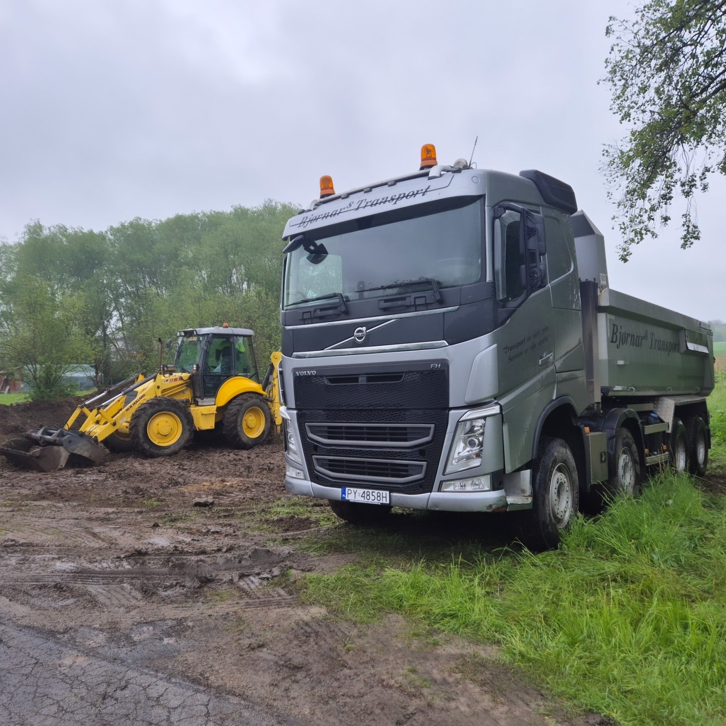 Żółta koparko-ładowarka New Holland w trakcie prac ziemnych obok szarej wywrotki Volvo z napisem 'Bjornar Transport' na tle zieleni i pochmurnego nieba.