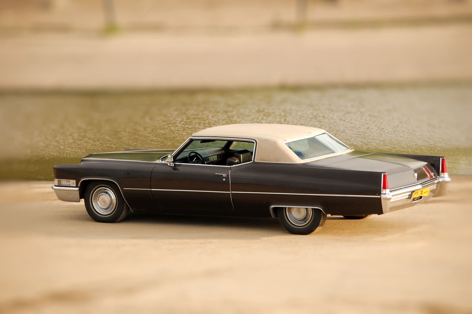 Ciemnobrązowy, klasyczny Cadillac z beżowym dachem winylowym, zaparkowany na piaszczystym brzegu z rozmytym widokiem na wodę w tle.