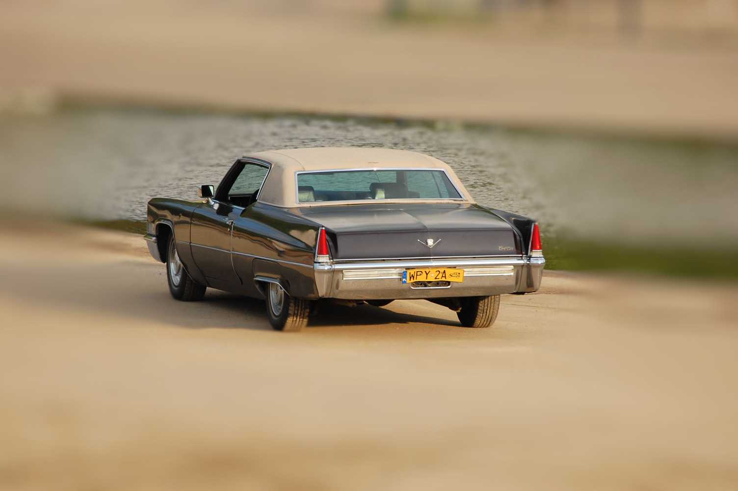 Czarny Cadillac Coupe Deville z beżowym dachem, widziany z tyłu na piaszczystym brzegu, z widoczną tablicą rejestracyjną WPY 2A.