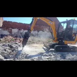 Żółta koparka JCB z młotem pneumatycznym kruszy betonowe elementy na tle ceglanego muru. Prace rozbiórkowe w słoneczny dzień.