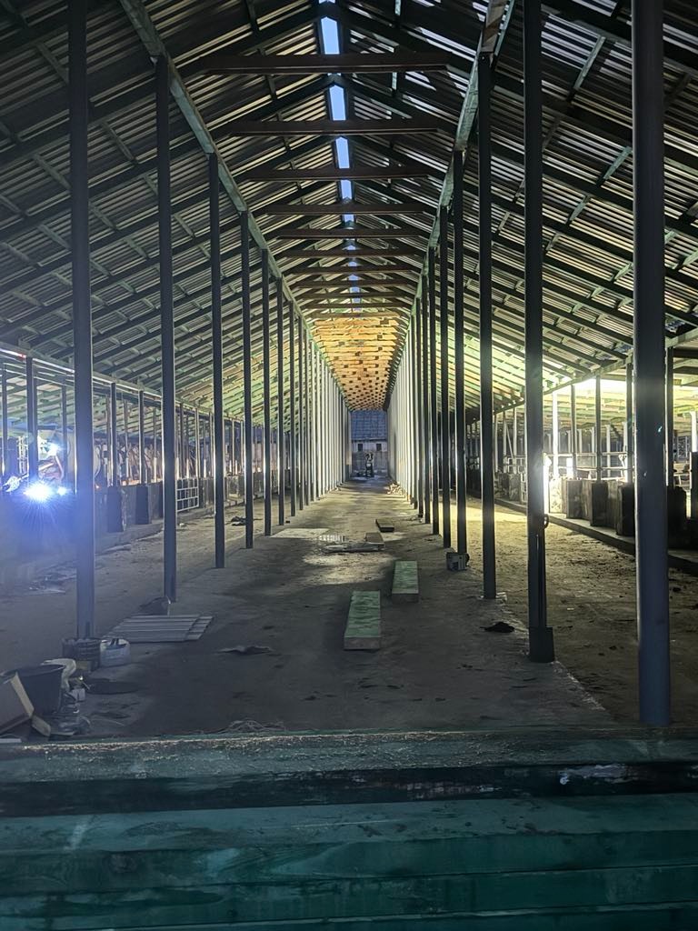 Wnętrze hali w trakcie budowy: metalowa konstrukcja dachu i słupy, surowa podłoga. Widoczne elementy oświetlenia i materiały budowlane w tle. Długi kadr perspektywiczny.