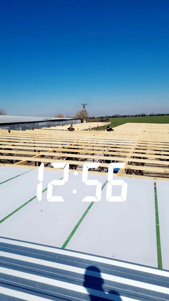 Konstrukcja dachu hali w trakcie budowy, widoczne drewniane elementy szkieletu i blaszane pokrycie, dwóch robotników w oddali, słoneczny dzień.