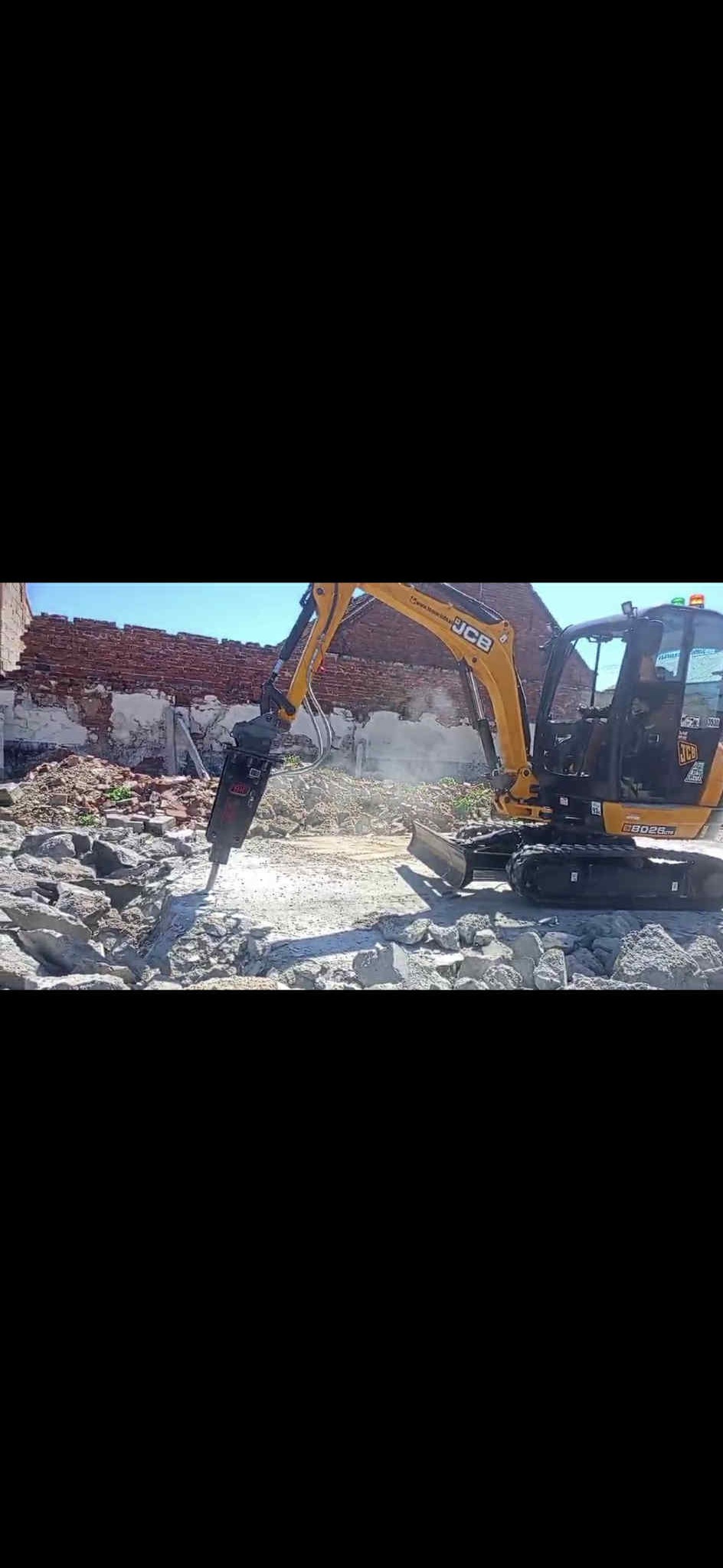 Żółta koparka JCB z młotem pneumatycznym kruszy betonowe elementy na tle ceglanego muru. Prace rozbiórkowe w słoneczny dzień.